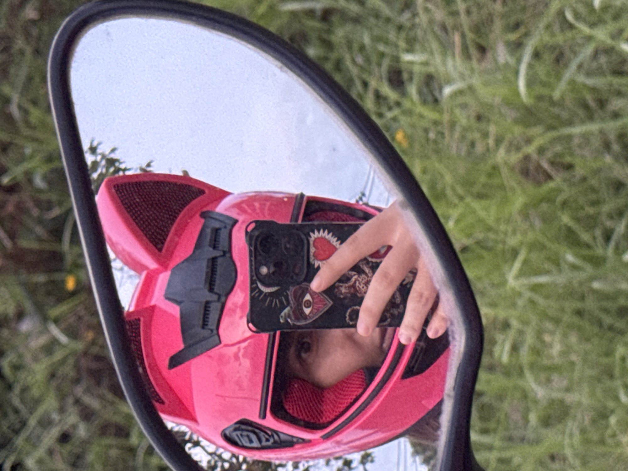 In a reflective mirror, a person wearing a bright pink helmet with cat-ear attachments is visible. The helmet has black accents and red interior padding. The person is holding a smartphone, also adorned with stickers of hearts and an eye, and is taking a selfie. The mirror's black frame is prominent, and the reflection shows a blurred background of green foliage and a pale sky, suggesting an outdoor environment. The location is Unknown, Unknown.
