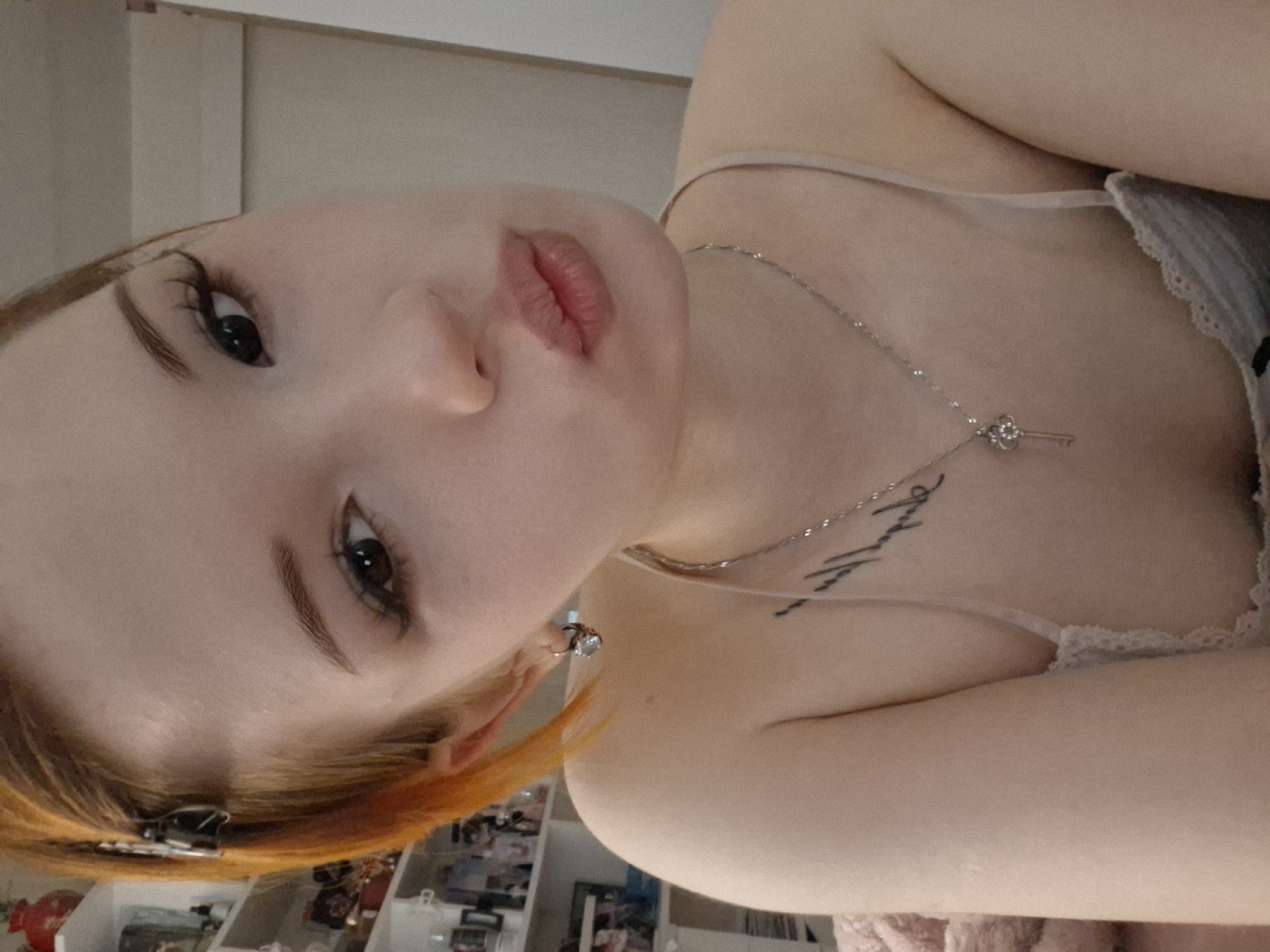 A close-up, rotated selfie shows a fair-skinned woman with red hair and dark eyes. She is wearing a light-colored camisole with lace trim and a silver necklace with a key pendant. A tattoo is visible on her chest. In the background, a shelf is filled with miscellaneous objects. The location in the city of Chaikovsky, Russia, is not discernible from the image.