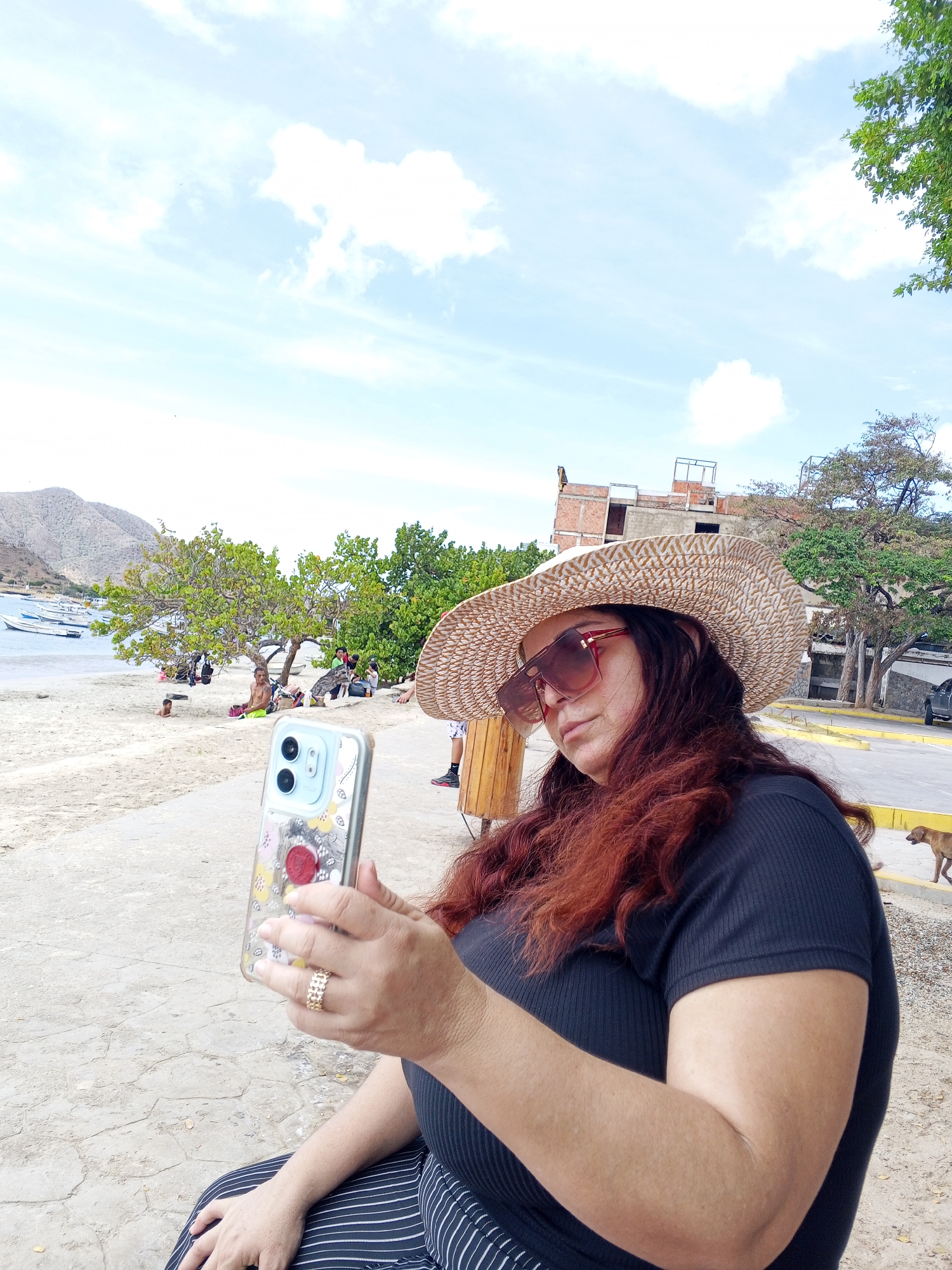 A woman with auburn hair, sunglasses, and a straw hat is holding a smartphone, possibly taking a selfie, outdoors. She is wearing a dark, short-sleeved top and striped pants. The location is near a beach, with a view of the ocean, mountains in the distance, and people relaxing on the sand. A building under construction is visible in the background. A dog is walking nearby. The city is Unknown, Unknown.