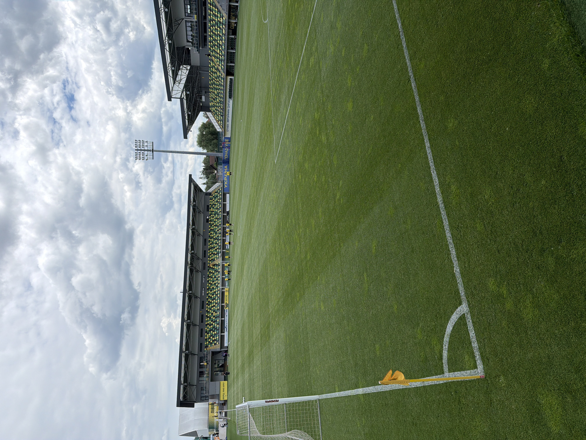 This is an outdoor, eye-level, wide-angle photograph of a green soccer field with stadium seating in the background. The field is well-maintained, with white lines marking the boundaries and a corner flag in the foreground left. There are several people on the field, some in yellow uniforms, appearing to be practicing. The stadium seating is predominantly green and yellow. Overhead, the sky is filled with dramatic clouds. The stadium appears to be located in Hnidyn, Ukraine, as indicated by the text on the banners in the background, which translate to "Ukraine is invincible."