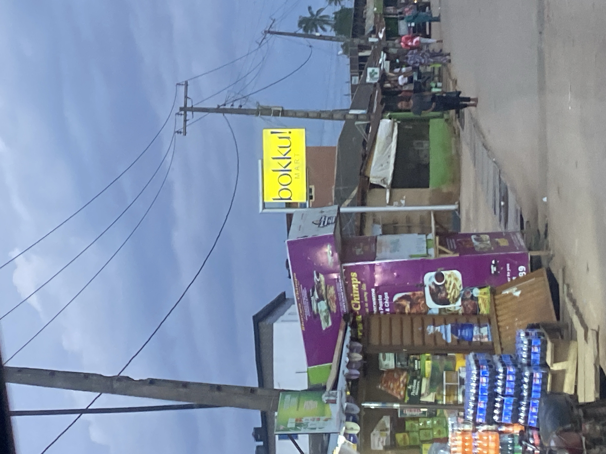 This is a medium-distance, eye-level, outdoor shot taken on a cloudy day in Ikorodu, Nigeria. The scene depicts a street view with several businesses and pedestrians. In the foreground, to the left, is a small shop stocked with bottled beverages, packaged snacks, and other goods, with a purple sign featuring images of food. Adjacent to this shop, a utility pole rises into the frame, crisscrossed with electrical wires. Further down the street, a bright yellow sign reads "bokku! MART". Several other low-lying structures, likely small businesses or market stalls, line the street. On the right side of the road, a few people are walking. One woman in a dark outfit is walking towards the camera, while others are visible further down the street. The overall impression is of a bustling, everyday street scene in a Nigerian city.