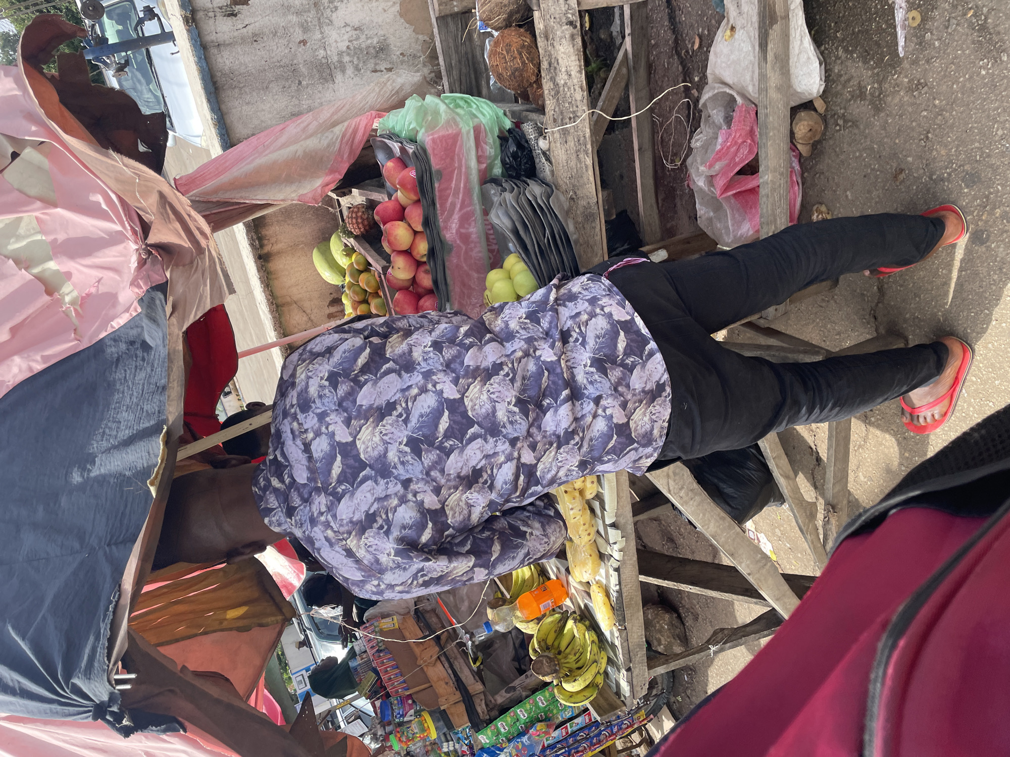 An outdoor fruit stand is depicted, situated on the side of a street in Jos, Nigeria. The stand is covered by a makeshift canopy constructed from various pieces of cloth, including pink, brown, and dark grey fabric. A man, viewed from behind, stands in front of the fruit display, wearing a patterned long-sleeved shirt and black trousers. He is wearing red flip-flops.
The fruit stand itself is a wooden structure laden with a variety of produce. Visible fruits include bunches of bananas, pineapples, apples, and coconuts. There are also some green watermelons or gourds. Several plastic bags, some containing items and others appearing empty, are present on and around the stand.
In the background, other individuals are partially visible, suggesting it is a busy area. A woman wearing a headscarf is seen at another stall to the left. Beyond the fruit stand, a light-colored vehicle is parked on a paved road. The ground appears to be a mix of asphalt and dirt. The overall impression is of a vibrant, open-air market scene.