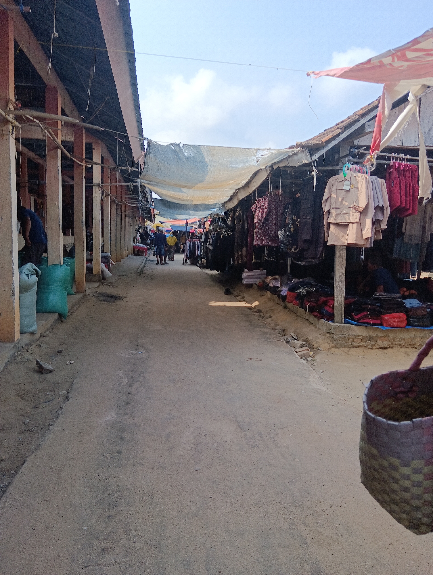 This image depicts a marketplace in Binakarsa, Indonesia. The scene is an outdoor market with stalls lining a dirt path. On the left, a structure with a dark roof supported by columns is visible, with several large, green sacks stacked near the entrance. Further down the path, clothing is displayed on racks and tables at various stalls. Some stalls have tarps or fabric coverings for shade. A few people are seen walking through the market and tending to the stalls. In the foreground on the right, a woven basket is partially visible. The sky is clear with some clouds. The overall impression is a busy, open-air market environment.