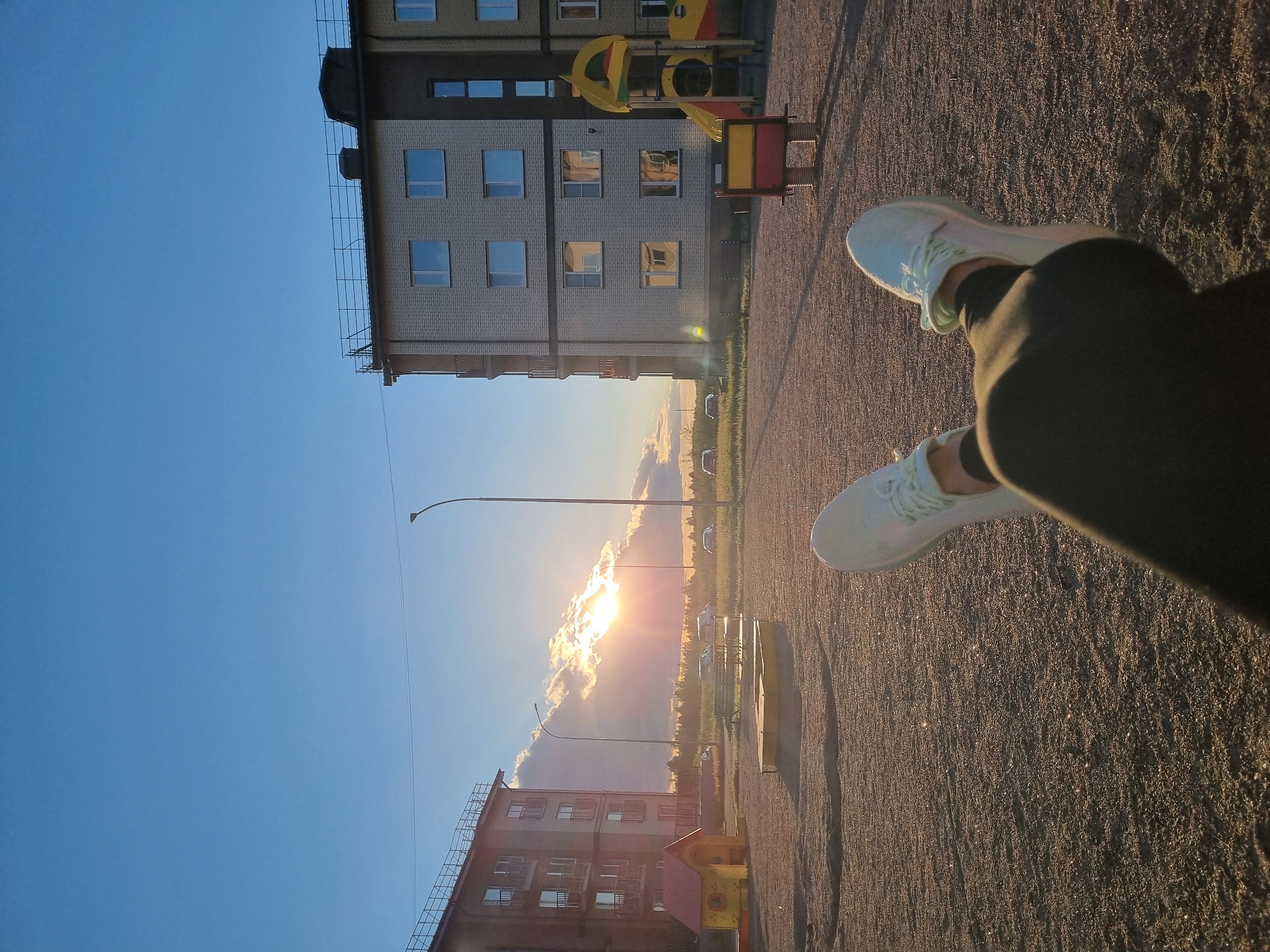 In an outdoor scene with a sandy ground, two legs with white sneakers are visible in the foreground, crossed at the ankles. The legs are clad in dark leggings. The background features a playground with a yellow and red slide, and two apartment buildings. A street lamp stands between the buildings. The sun is setting, casting a warm glow on the scene and creating lens flare. Several cars are parked in the distance, and a line of trees borders the horizon. The sky is a clear blue with a few clouds illuminated by the setting sun.
