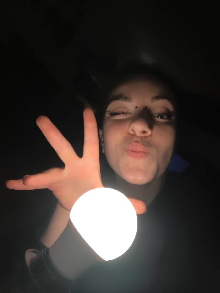A young woman with dark hair poses in a dimly lit area, holding a bright, spherical light in her outstretched hand. She is winking one eye and puckering her lips. The light illuminates her face, revealing eyebrow piercings and dark eye makeup. Her other hand is open, fingers spread. The background is mostly black, suggesting nighttime or a dark room.