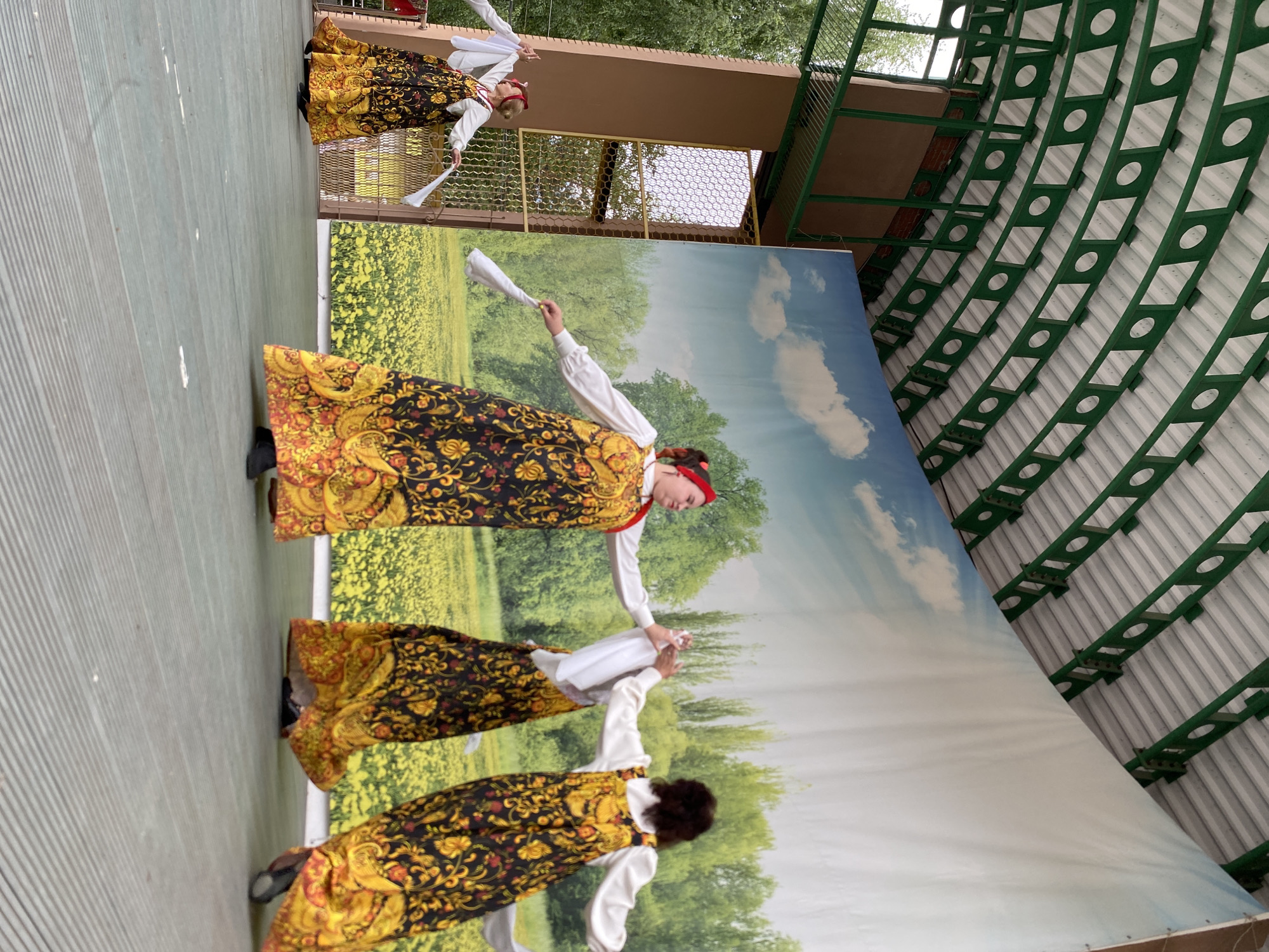 Four women in traditional Russian folk costumes are performing a dance on an outdoor stage. The costumes feature long, flowing skirts with intricate yellow and red floral patterns on a black background, paired with white blouses and red headscarves. Each dancer holds a white scarf, manipulating it as part of their choreography.
The stage is covered with a light green surface. Behind the dancers is a large backdrop depicting a sunny landscape with a field of yellow flowers under a bright blue sky with white clouds, and trees in the distance. Above the stage, a green, arched metal structure with circular cutouts supports a white ceiling. To the left of the stage, a brown structure with a latticed window is visible, with trees and foliage outside.
This scene is likely taking place at an outdoor venue in the city of Chaikovsky, Russia, possibly during a cultural festival or performance. The setting suggests an open-air theater or a similar public space designed for entertainment.