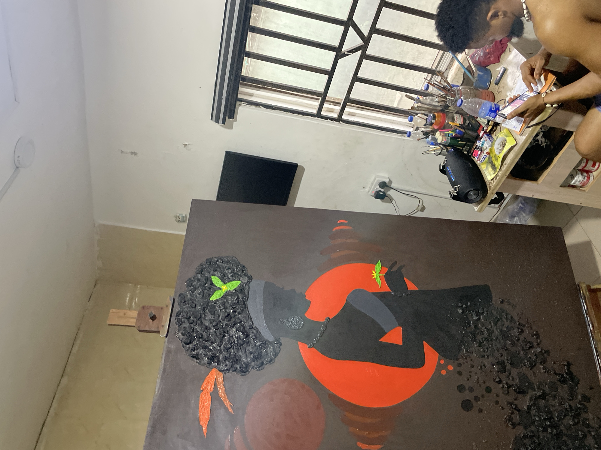 A medium-shot, interior view, shows an individual working on a painting, possibly in a studio space located in Ado, Nigeria. The individual, a dark-skinned man with short black hair, is seated at a table and appears to be consulting materials, possibly a phone or a book. The table is cluttered with art supplies, including paintbrushes, paint containers, and water bottles. Adjacent to the table is a black speaker.
The focal point of the scene is a large canvas displaying a painting depicting a woman's silhouette against a red circular backdrop, possibly representing a sun or moon. The woman has an Afro hairstyle with green leaf embellishments. Orange paint accents are also present. The painting is set up on an easel.
In the background, a window with metal bars is visible, along with a black television screen mounted on the wall. The walls are painted white, with some visible imperfections or marks. The lighting is a mix of natural light coming through the window and artificial light from an unseen source. The overall impression is of a creative and lived-in workspace.