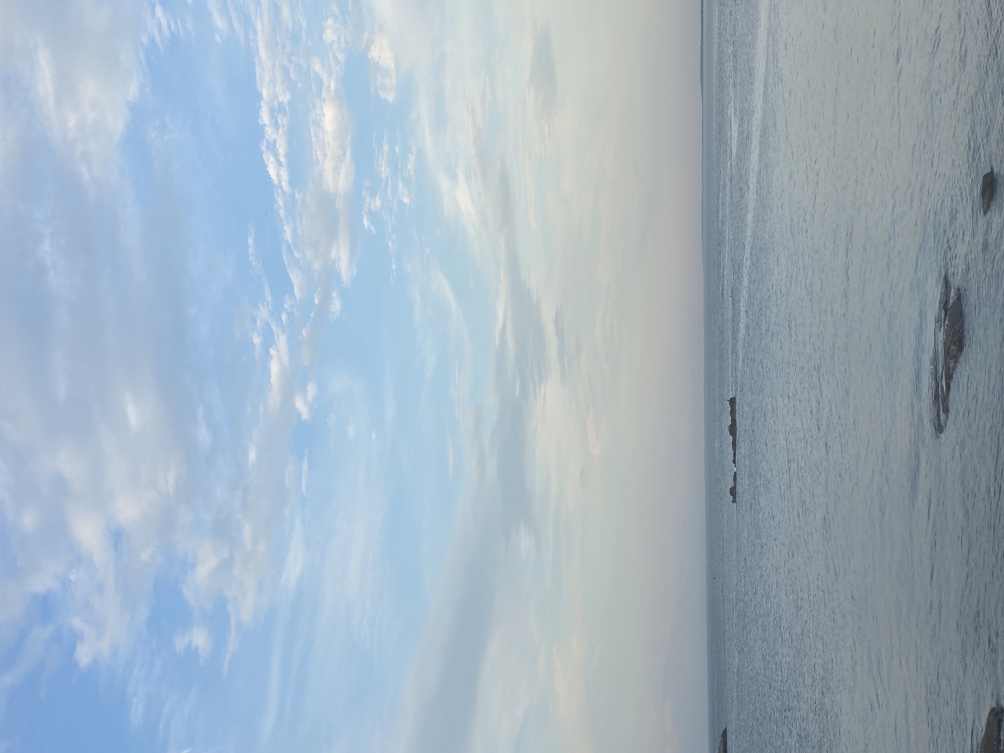 The image depicts a view of the Long Island Sound in the City of New Rochelle, United States of America. The left side shows a bright blue sky with scattered white clouds. The right side features the calm, gray water of the Long Island Sound with rocks protruding near the shoreline. There are small, indistinct objects in the water further from the shore. The horizon line is visible where the sky and water meet.