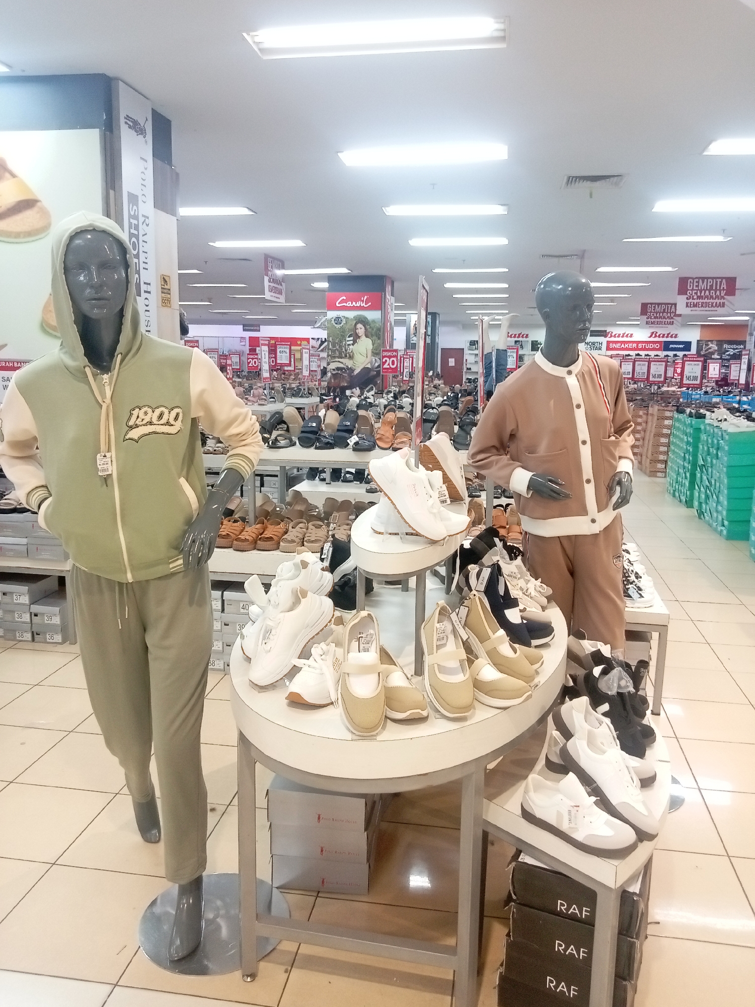 This image shows a retail store in Batam, Indonesia, likely a shoe store within a larger department store, possibly Nagoya Hill Shopping Mall, given the common store types found there. The foreground features two mannequins displaying casual apparel and footwear. The left mannequin is a male model dressed in a light green hooded jacket with "1900" emblazoned on the chest, paired with matching light green trousers. A display of white sneakers is arranged around its base. To the right, another male mannequin is dressed in a light brown jacket with white trim and matching trousers, also showcasing footwear.
The store is well-lit with fluorescent ceiling lights. In the background, numerous shelves are filled with various styles of shoes, including sandals, heels, and athletic shoes, catering to a wide range of customers. Several promotional signs, some with discounts like "DISKON 20%" and "DISKON 65%", are visible, indicating ongoing sales. The floor is tiled with a grid pattern. The overall impression is that of a busy and well-stocked retail environment.