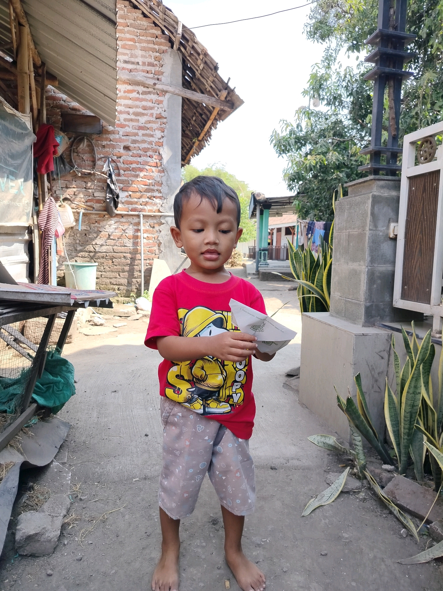 In Beloh, Indonesia, a young boy stands in a dirt courtyard. He is wearing a red t-shirt with a yellow cartoon character and patterned shorts. The boy is holding a folded piece of paper. Behind him, a brick building with a corrugated metal roof is visible. To the right, a tall, dark metal structure stands next to a concrete pillar and a white fence with decorative elements. Several snake plants are growing near the pillar. The overall environment appears to be an outdoor residential area.