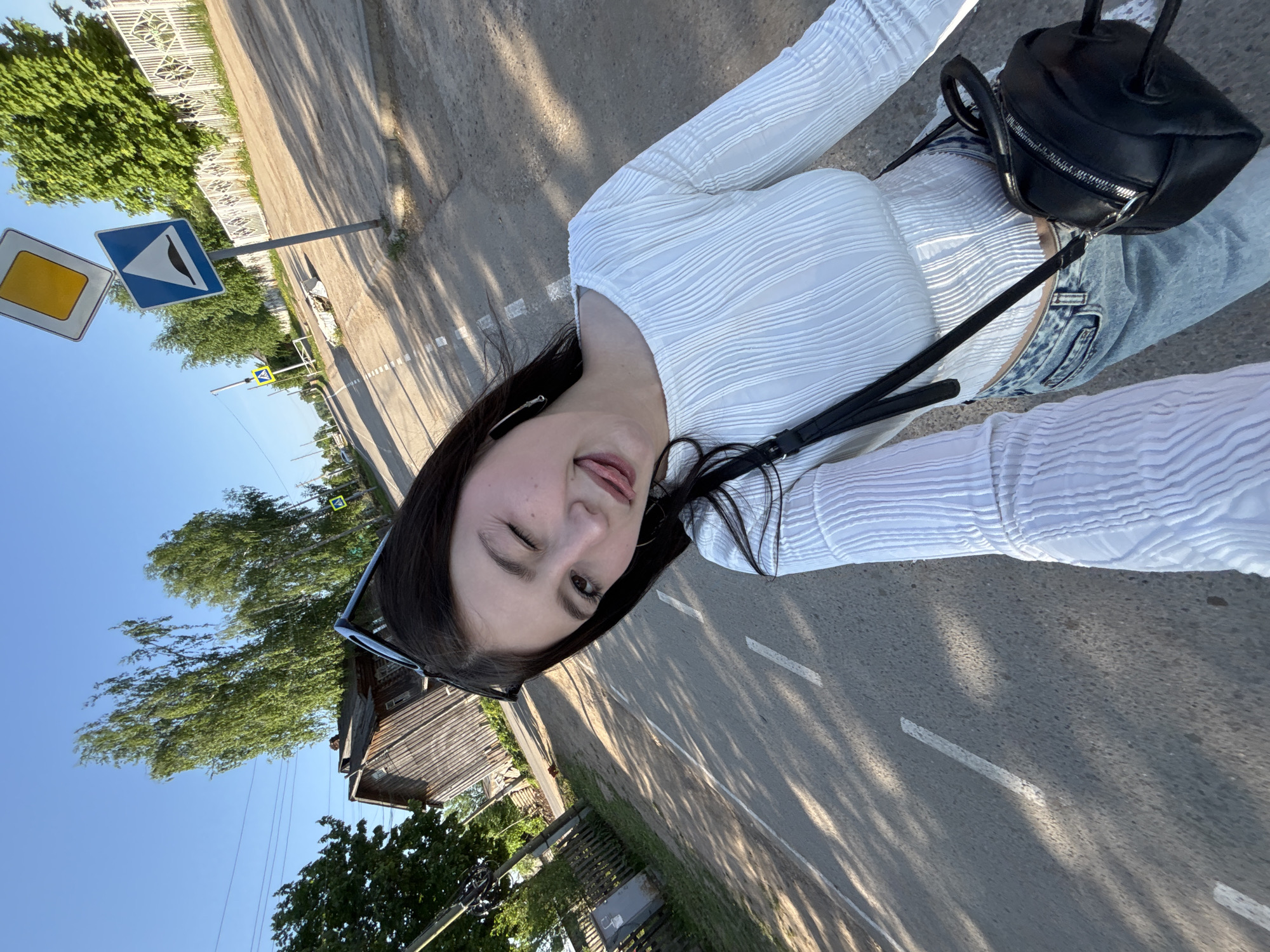 A young woman stands on an asphalt street in Фоки, Russia, taking a selfie. She is wearing a white long-sleeved top and light-wash jeans, with a small black purse slung over her shoulder. She is winking at the camera, with sunglasses perched on her head.
The setting is a rural street with wooden houses and greenery visible on either side. Road signs are present, indicating directions and traffic regulations. The road has white painted lines. Shadows of trees are cast across the asphalt. The sky is a clear blue.