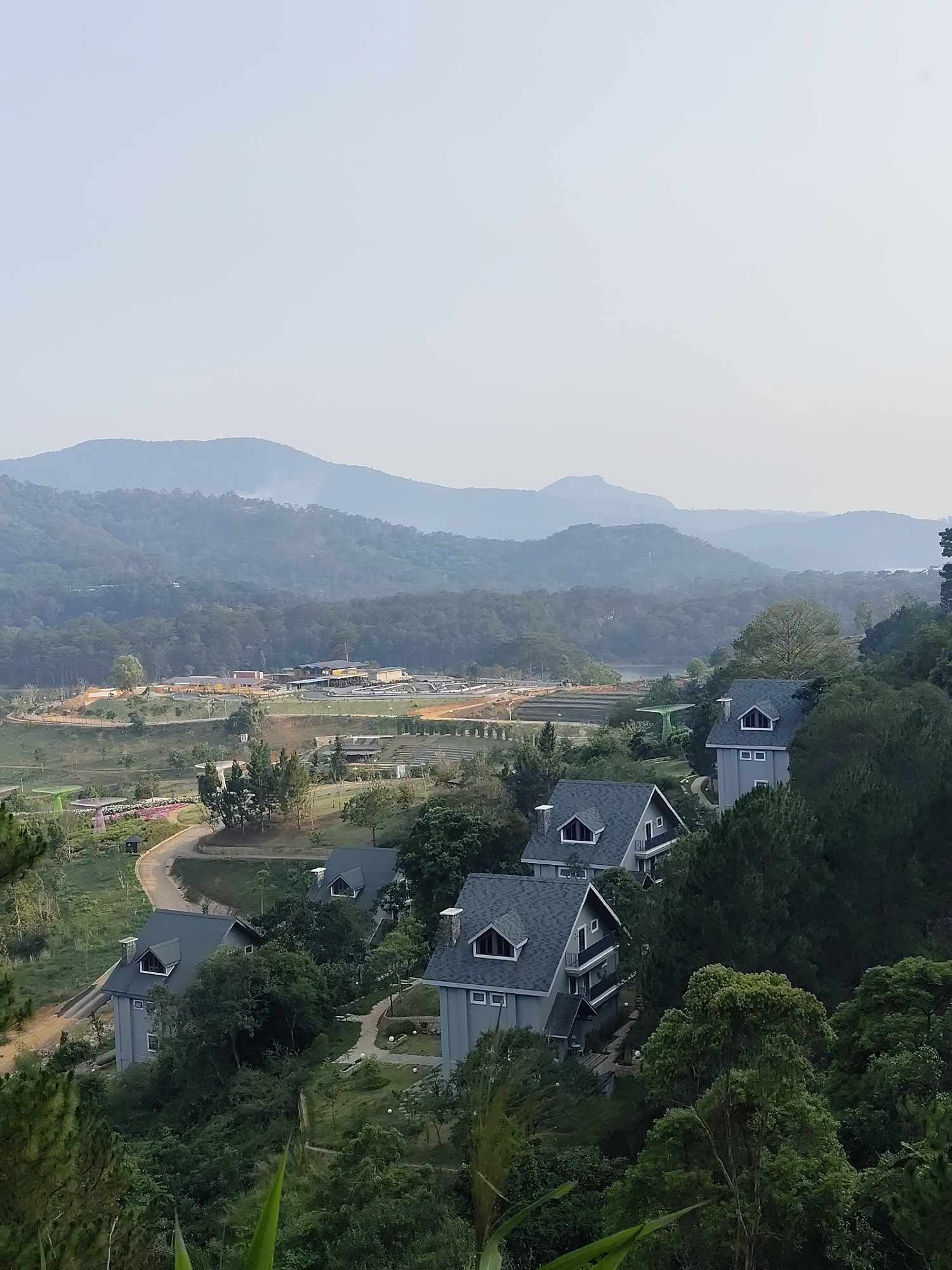 The image depicts a hilly landscape in Da Lat, Vietnam. In the foreground, several modern, grey-colored houses with dark roofs are nestled among lush green trees. A winding road leads from the houses towards a more developed area in the mid-ground, featuring buildings, open spaces, and what appears to be a small pond. In the background, layered mountains fade into a hazy sky. No people are visible in the image.