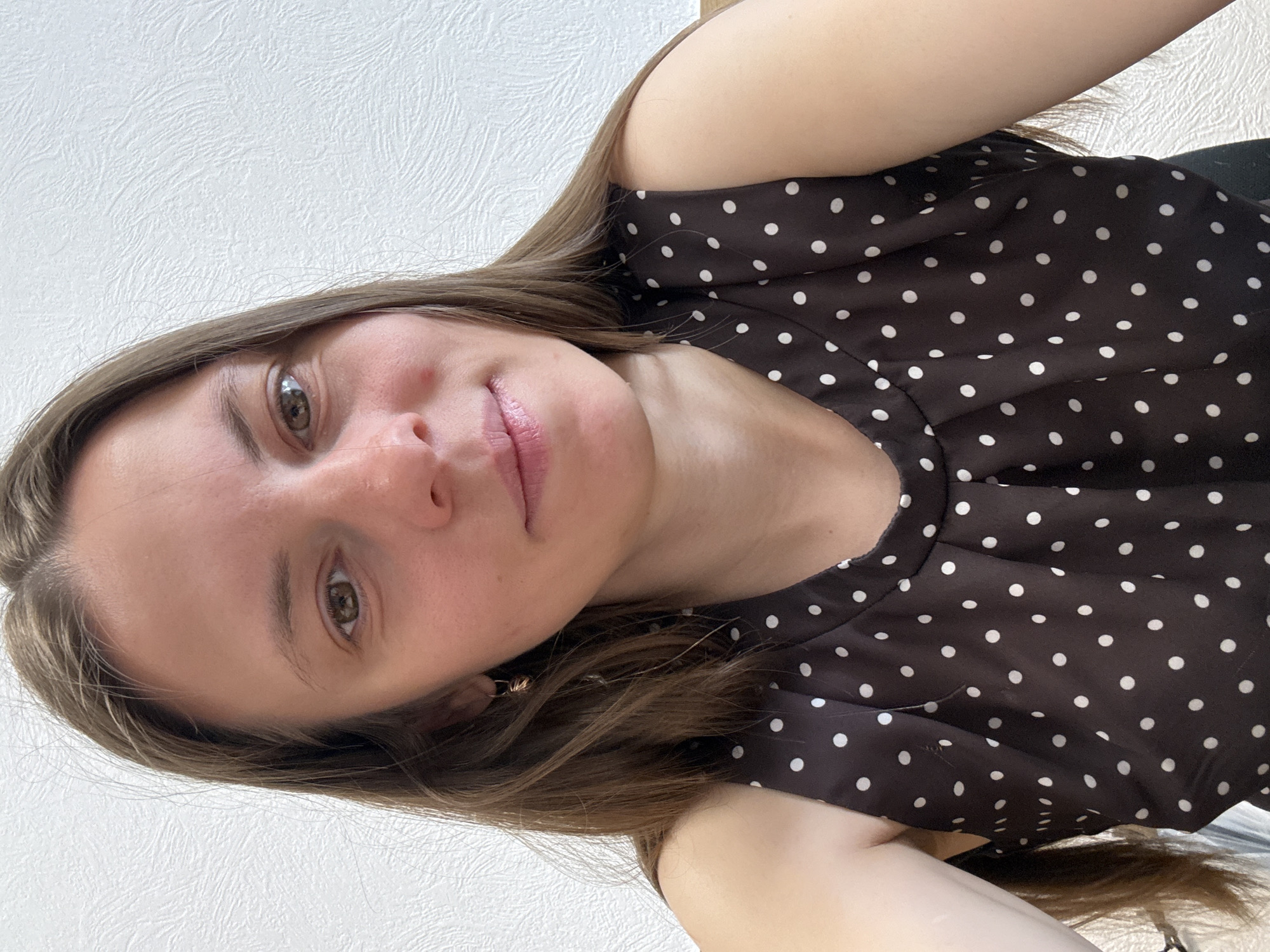 A woman is photographed in a close-up selfie. She has light brown hair, fair skin, and green eyes. She is wearing a dark brown, polka-dotted top. The background appears to be a white wall. The location in Chaikovsky, Russia is not identifiable from the given image.