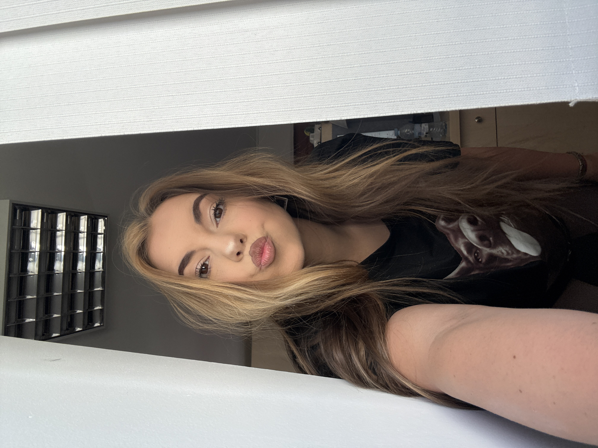 A young woman with blonde and brown hair is centered in the frame, looking directly at the camera with a slightly pouting expression. She is wearing a black t-shirt with a white print on it. The background is partially obscured by white Venetian blinds, with a window to the left showing a view into a darker space. There are items visible on a shelf above her. The location is not discernible from the image. The city of Sofia, Bulgaria, cannot be confirmed as the location.
