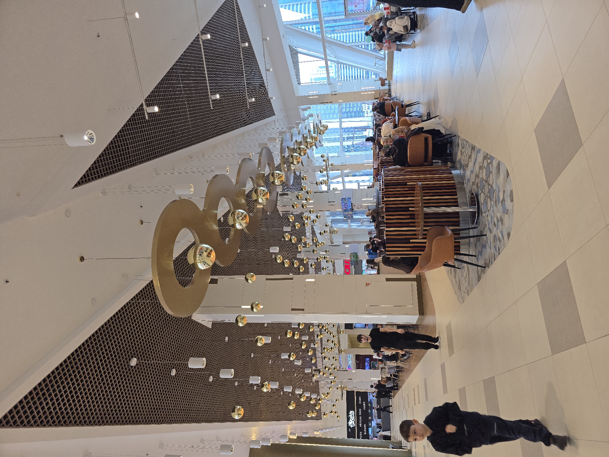 The image depicts the interior of the Grozny Mall in Grozny, Russia. The mall has a modern architectural design with a high ceiling featuring decorative geometric elements, including triangle-shaped lattice panels and a series of large, golden ring-shaped light fixtures with spherical lamps suspended from them. The floor is tiled with light-colored squares and rectangular patterns. People are scattered throughout the scene; some are seated at tables in a cafe or lounge area, others are walking through the mall. There is a mix of adults and children present, with some of the children lying on the floor. There are various storefronts and signs visible in the background. Natural light enters the mall through the glass ceiling and window structures.