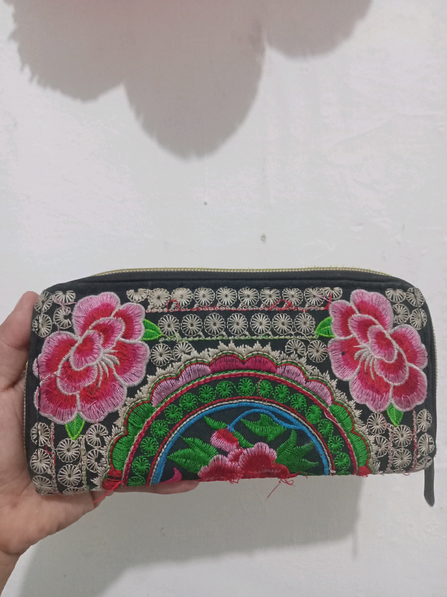 A person's hand is holding a black zippered wallet with intricate floral embroidery in vibrant pink, green, and blue. The wallet is positioned horizontally. The background is a plain, white wall with a shadow cast on it from an unseen object, possibly the person holding the wallet or another object. The image appears to have been taken indoors. There is no information available to determine the location in Sukomananti, Indonesia.
