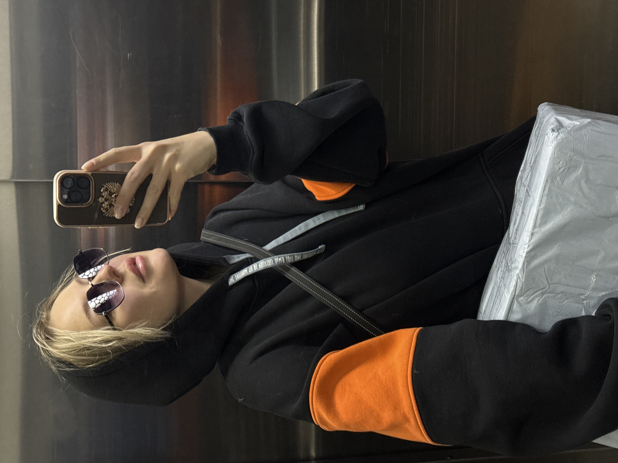 A person with fair skin and blonde hair wearing sunglasses is taking a selfie with a smartphone. The phone has a dark case with a gold-colored emblem. The person is wearing a black hoodie with orange accents on the sleeves and a gray drawstring. The backdrop appears to be a metallic or stainless steel surface, possibly in an elevator. The image appears to be taken sideways. There is a wrapped package near the bottom right of the frame. There is no information in the image that suggests it was taken in Krasnoyarsk, Russia.