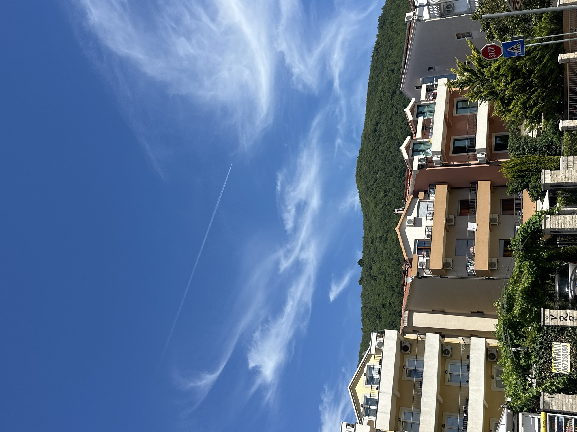 In Budva, Montenegro, a clear blue sky with wispy cirrus clouds stretches above a dense, green, forested mountain. In the foreground, several multi-story buildings are visible, featuring balconies with railings and visible air conditioning units. The buildings are a mix of yellow, beige, and pink hues. Lush green vines are growing over an archway in front of one of the buildings, and a sign reads "Apartmani" followed by a phone number. A silver SUV is parked behind a stone wall. Further to the right, a red octagonal stop sign and a pedestrian crossing sign are mounted on poles next to a conical evergreen tree.