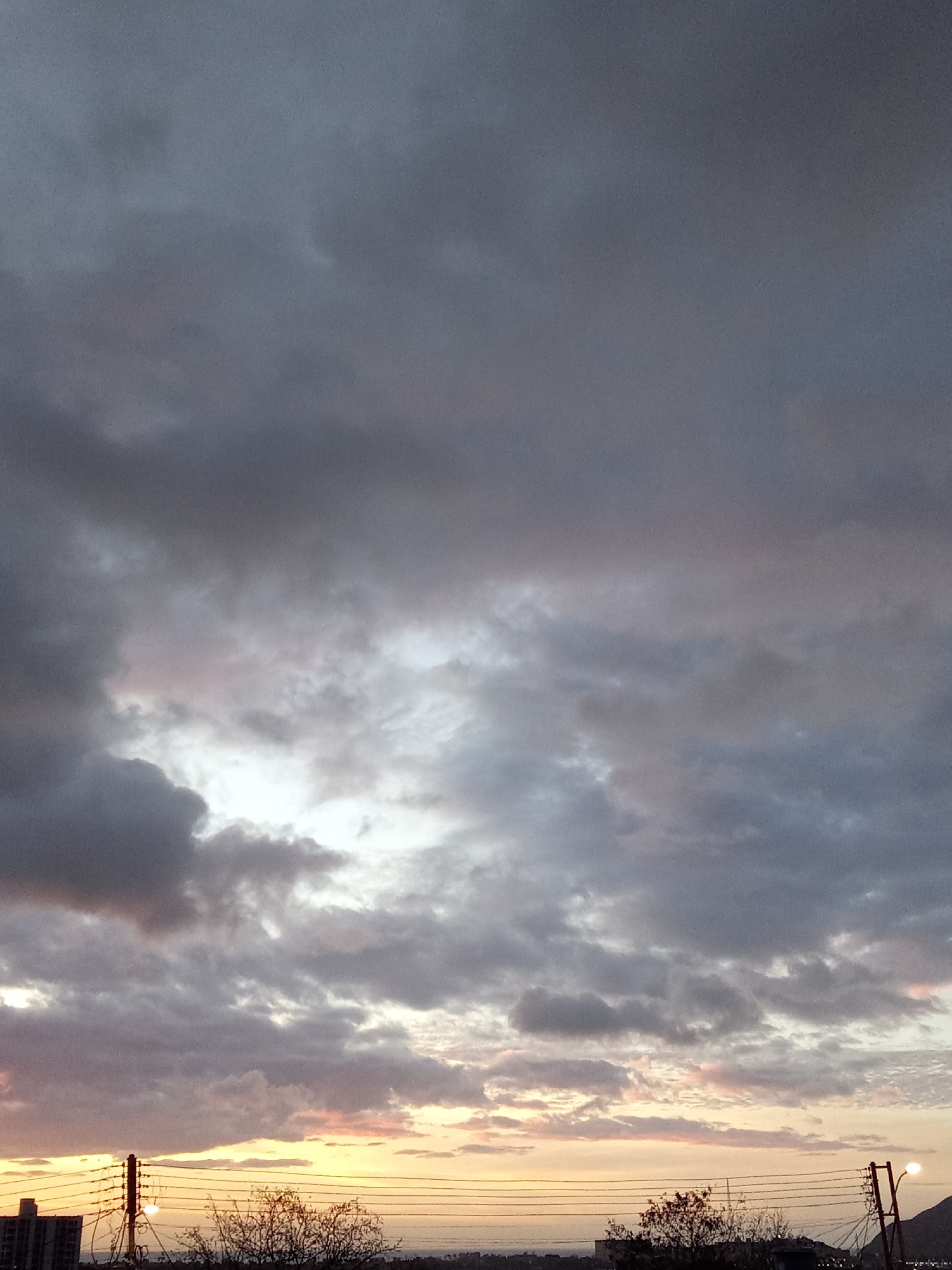 The image captures a sky filled with gray and white clouds, with a visible break in the clouds allowing light to shine through. The bottom of the frame shows a distant cityscape at the city of Juan Griego, Venezuela. Utility poles and wires are visible in the foreground. Silhouetted trees and buildings appear along the horizon where the sky meets the land, indicating a sunset or sunrise. The light behind the clouds hints at pink and orange hues.