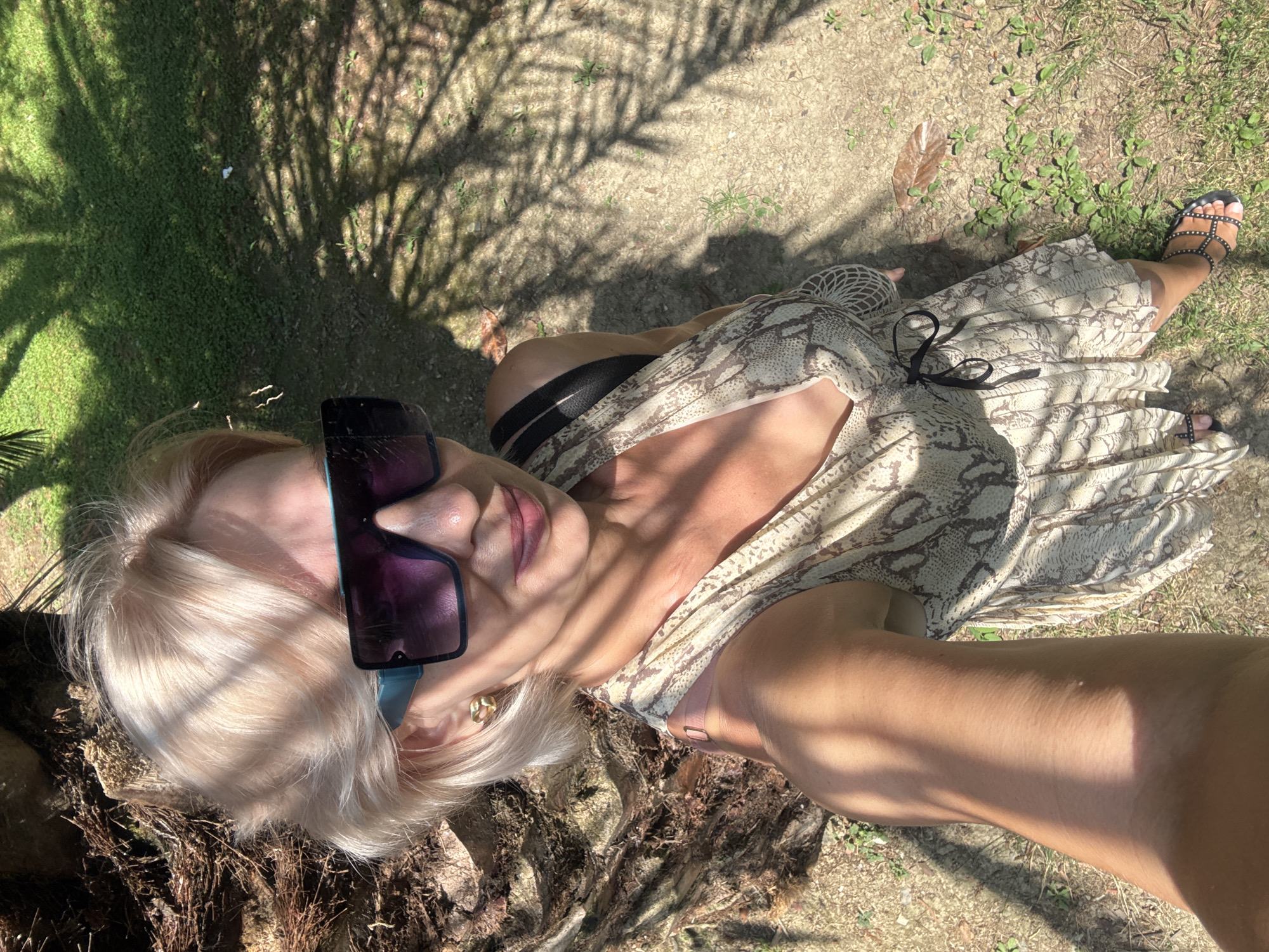 A woman with blonde hair and large sunglasses takes a selfie outdoors. She is wearing a light-colored dress with a snake-like pattern and black strappy sandals. She stands next to a palm tree, with shadows from palm fronds cast across the ground. The ground appears to be a mixture of dirt and grass. This image is located in Sochi, Russia, a city known for its subtropical climate and palm trees, often found in parks and along the coast.