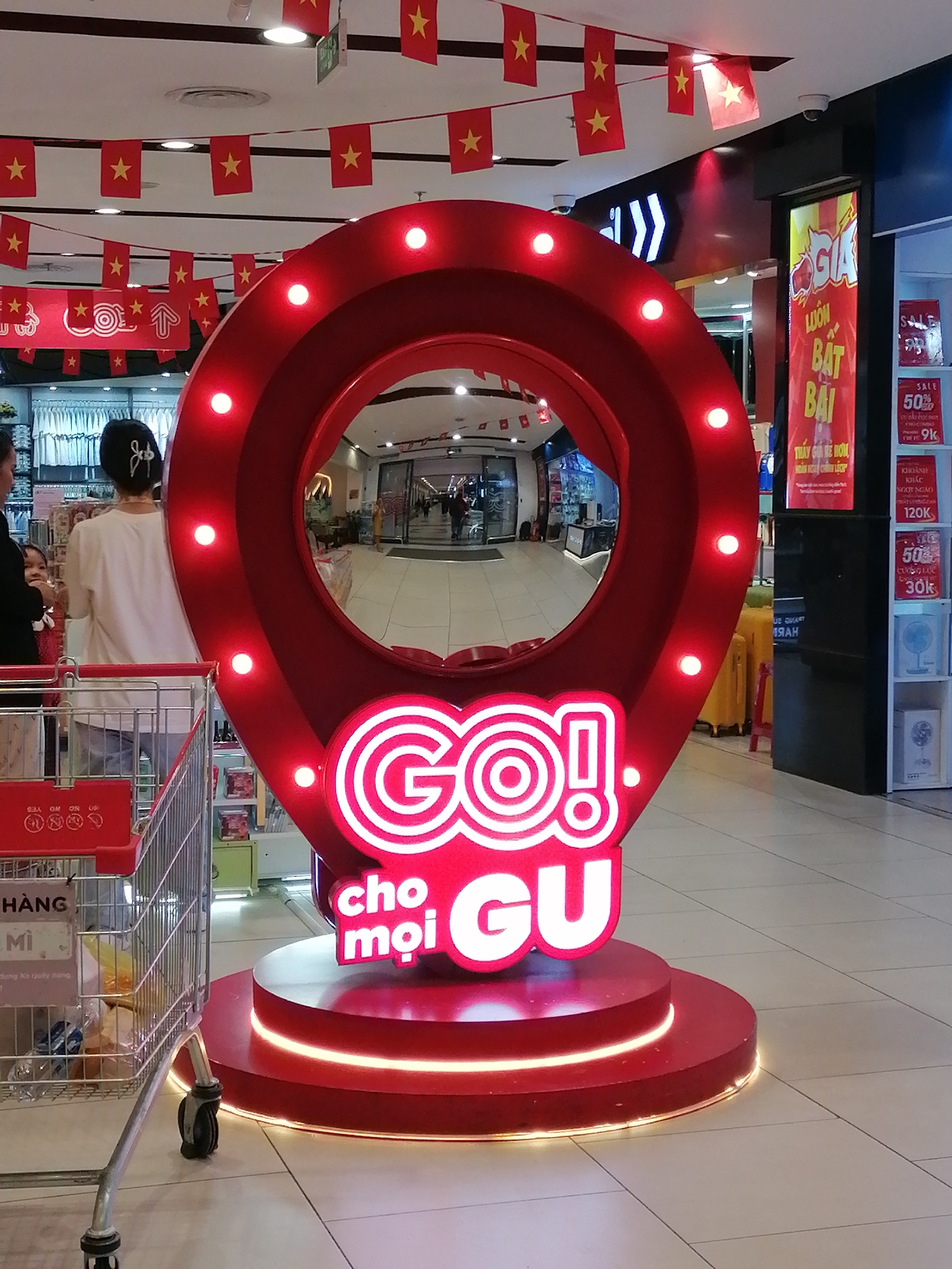 The image shows a brightly lit "GO!" promotional display, shaped like a location pin, in what appears to be a shopping mall in Da Lat, Vietnam. The display is red with white neon lettering. The text "cho moi GU" is displayed under "GO!" The display is on a multi-tiered circular red base with integrated lighting.
In the background, a woman with a hair accessory is seen with a shopping cart. Banners of red flags with yellow stars (Vietnamese flag) are hanging from the ceiling. A store sign with sale information is visible on the right side. People are walking in the background through the mall.