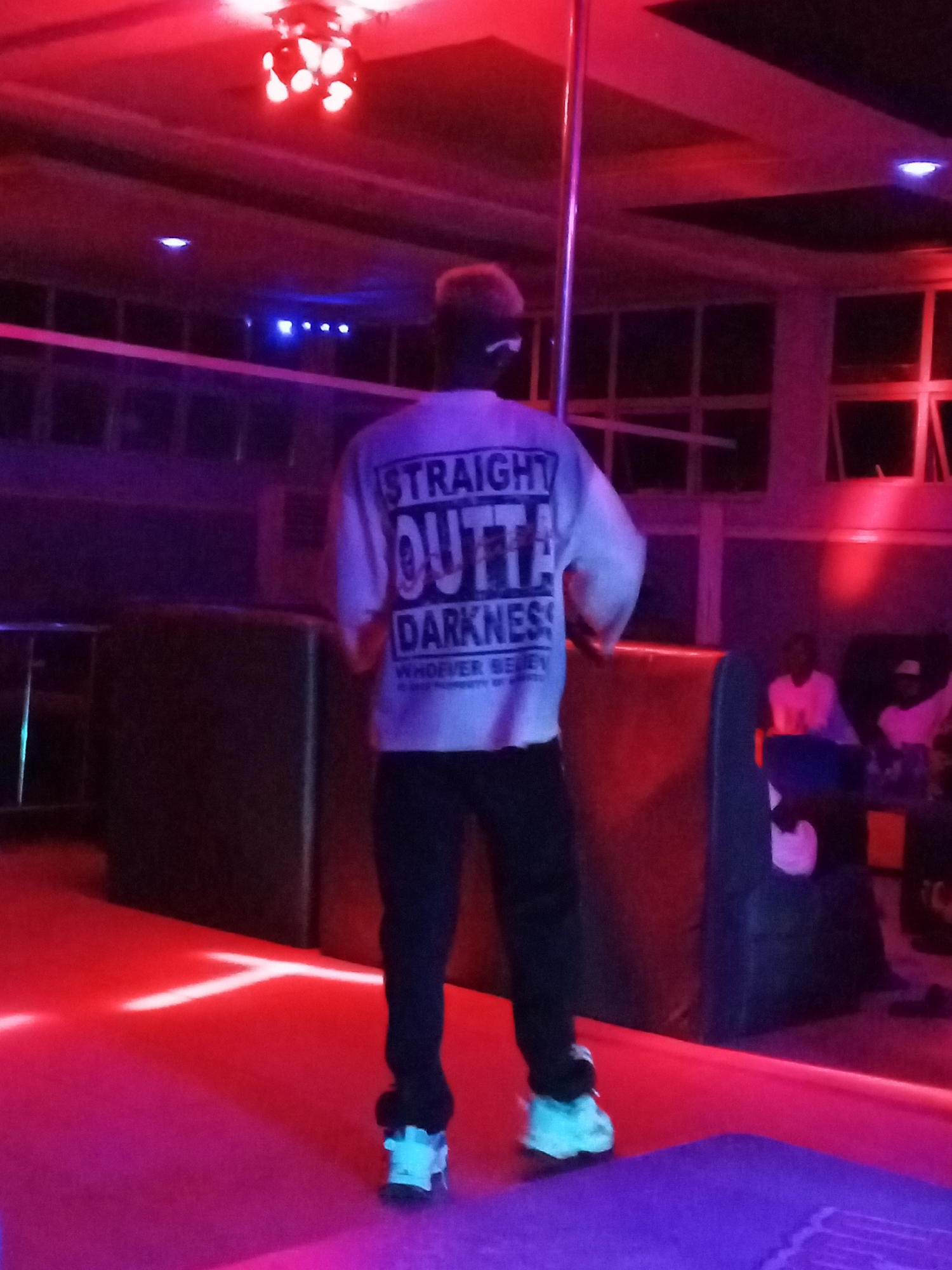 A person in a white shirt that says "STRAIGHT OUTTA DARKNESS" is standing on a stage in a dimly lit room with red and purple lighting. The person is wearing black pants and blue sneakers. There is a pole on the stage and people are seated in the background. The location is in the city of Unknown, Unknown.