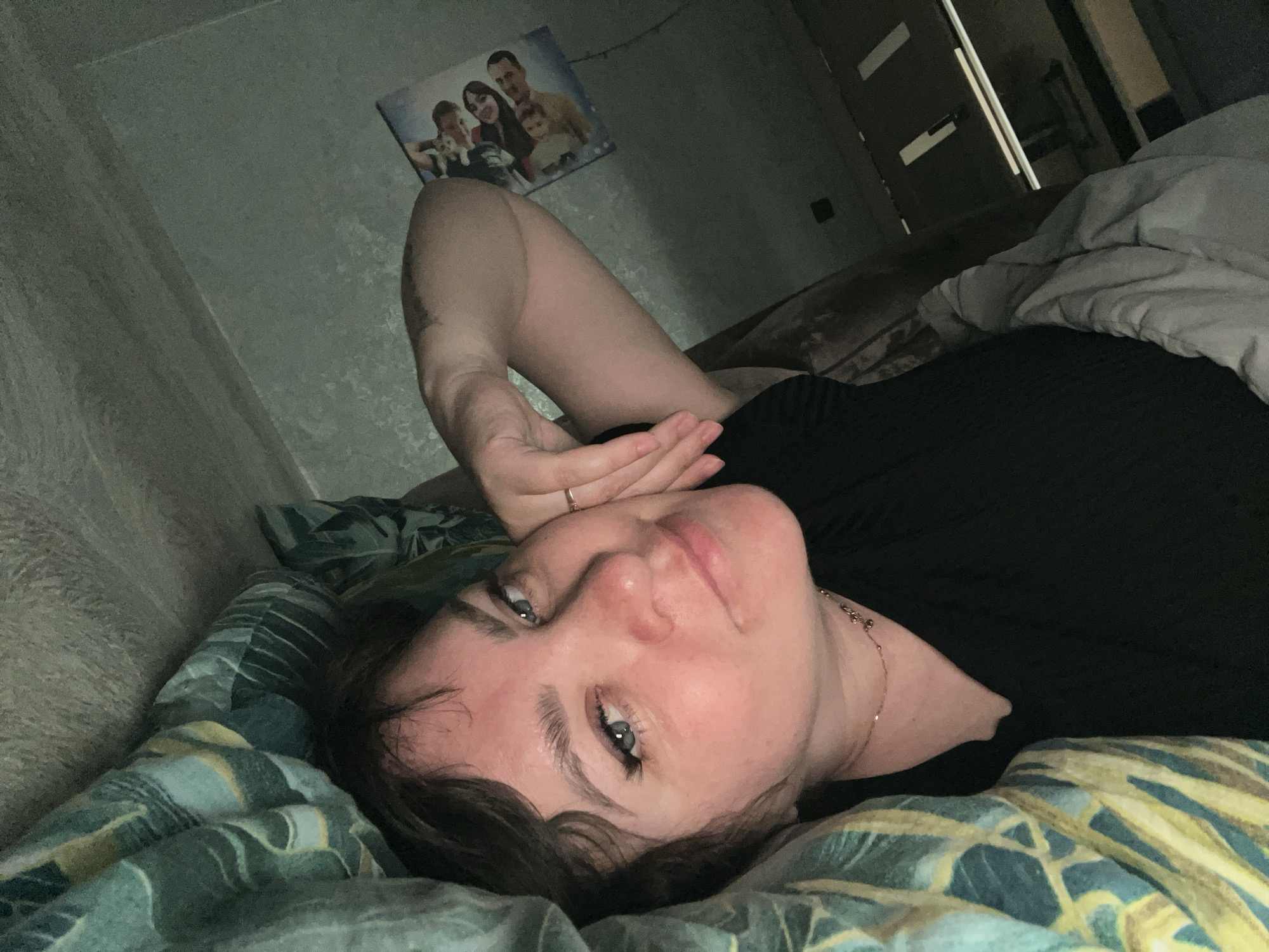 A person is lying on a bed covered with a patterned blue, green, and yellow sheet. The person is wearing a black top and has dark hair and fair skin. Their right arm is raised, and their hand is near their face. They are looking at the camera. A framed family photo hangs on the wall above the bed. A closed dark-colored door is visible in the background. The photograph was taken in an indoor location. There is no indication this image was taken in the city of Noviy, Russia.