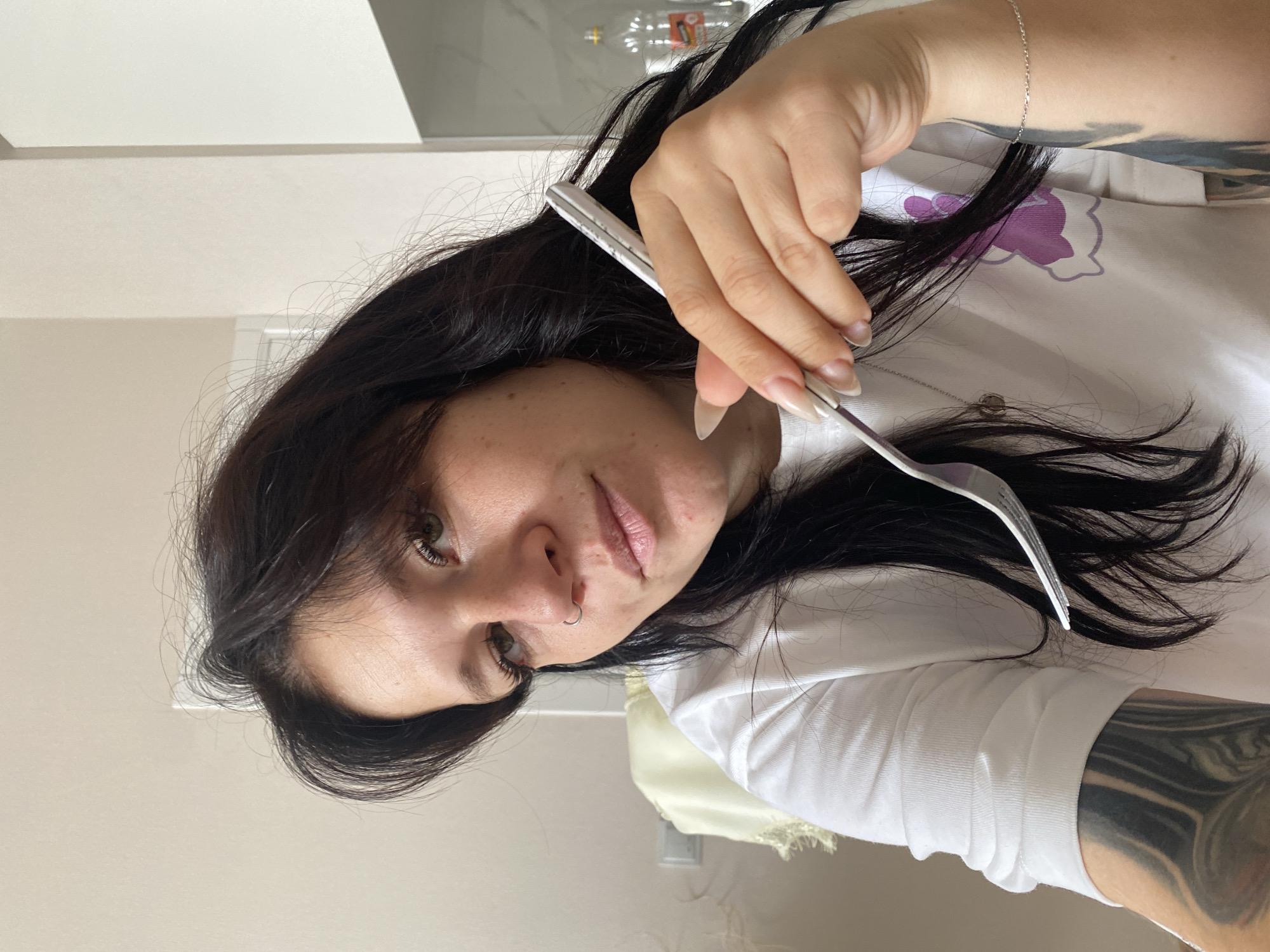 In the city of Yanino-1, Russia, a young woman with dark hair, adorned with a nose ring and long fingernails, is captured in a close-up portrait. She is wearing a white long-sleeved shirt with a purple graphic design. Her right hand holds a silver fork, with which she appears to be interacting with her hair. The background is blurred, suggesting an indoor setting, possibly a kitchen or dining area, with a hint of white cabinetry and a glimpse of a bottle on a shelf. The woman has tattoos on her arm.