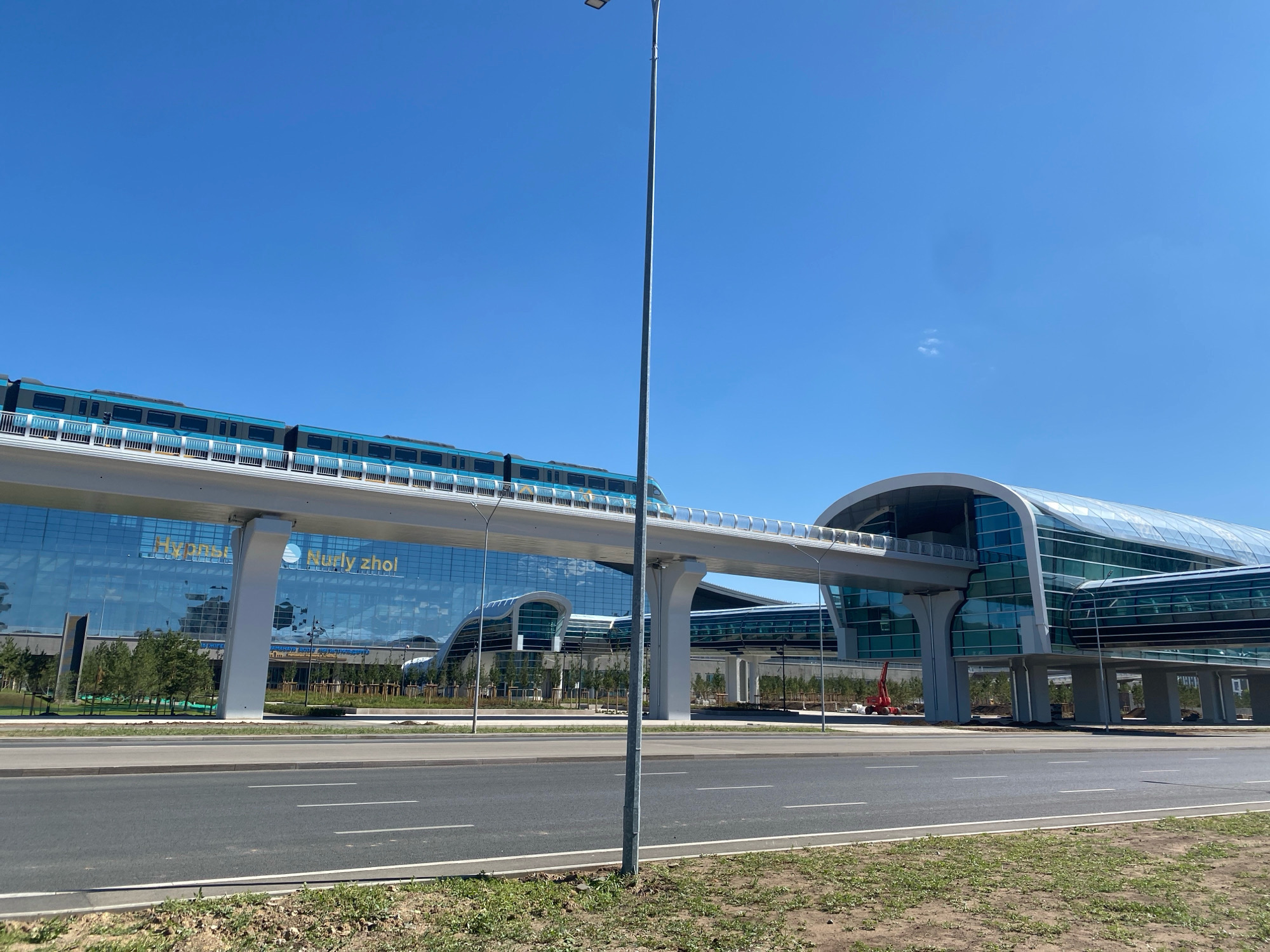 A light blue and gray monorail train travels along an elevated track in front of a modern building with a glass facade. The building is located in Astana, Kazakhstan, and appears to be a transportation hub or station. The architecture of the building is sleek and contemporary, with curved lines and large expanses of glass that reflect the sky and surrounding trees. The monorail is a prominent feature, indicating a public transportation system. The scene is set against a clear blue sky, suggesting a sunny day. The foreground shows a paved road with white lane markings, and a grassy verge. The overall impression is one of a well-developed urban environment with modern infrastructure.
