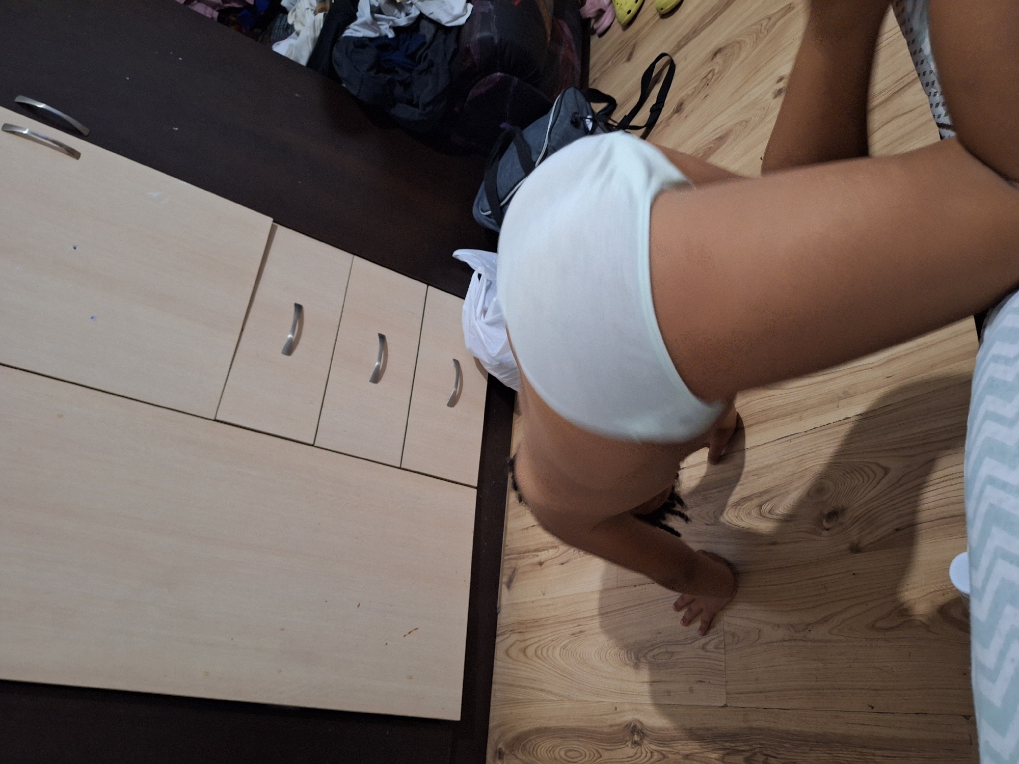 The image shows a person wearing white underwear in a home setting. The person is in a crawling or squatting position, with their back to the viewer. The background includes a wooden floor, a dresser with drawers, and some laundry and bags on a dark surface. The lighting suggests it is daytime indoors. There is no visible text.