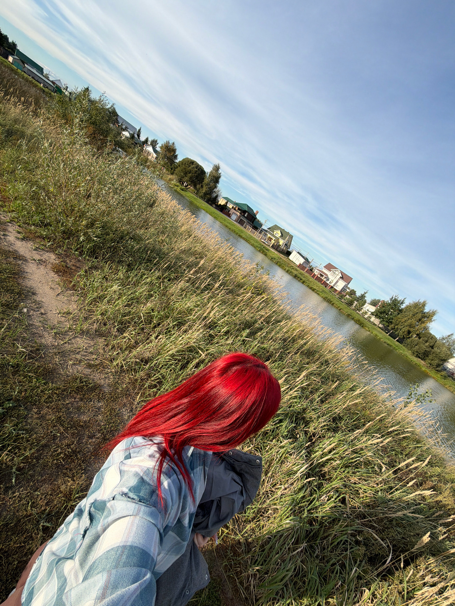 In the city of Uzuykovo, Russia, a person with bright red hair is captured in a selfie from a high angle. They are wearing a plaid shirt over a dark jacket and are positioned on a sandy path bordered by tall, dry grass. To the right of the path is a calm body of water, likely a river or lake, reflecting the clear blue sky with sparse white clouds. On the opposite bank of the water, several residential houses with green and brown roofs are visible, nestled among trees. The scene is illuminated by sunlight, casting shadows on the ground and grass.