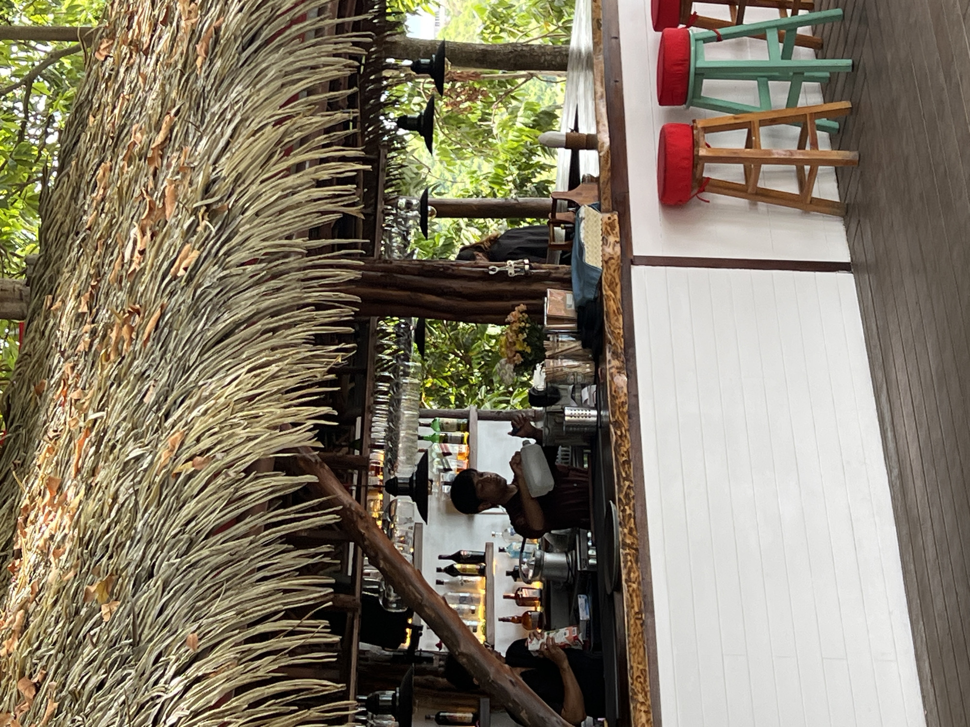 The image depicts an open-air bar area with a thatched roof, likely located at a beachside resort in Baan Chaweng Noi, Thailand. A wooden bar counter spans the foreground, with white wooden paneling beneath it. Three bar stools are visible: one light wood with a red cushion, a teal stool with a red cushion, and a third partially visible stool with a red cushion.
Behind the counter, two individuals are working. One, wearing a maroon shirt, is pouring liquid from a clear jug into a stainless steel container. The other individual, wearing a black shirt, is handling a white bag. Shelves behind them are stocked with bottles and glassware.
The environment surrounding the bar is lush and green, suggesting a tropical setting with abundant foliage. Natural light filters through the thatched roof and the surrounding vegetation. The overall atmosphere suggests a relaxed, tropical establishment.