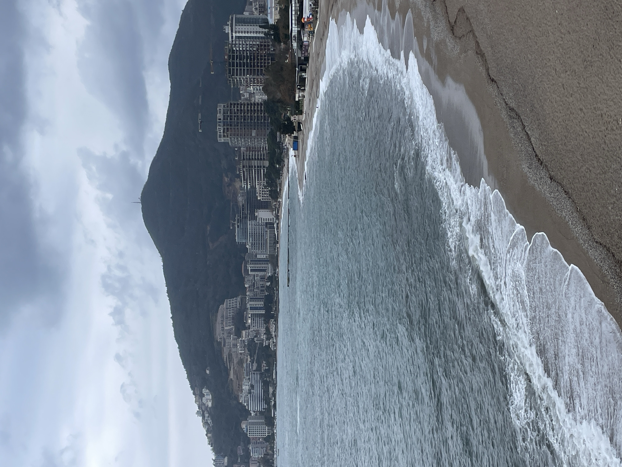 The image displays a panoramic coastal view of Budva, Montenegro, under an overcast sky.
In the foreground, a sandy beach, predominantly light brown, extends along a gentle curve. White-capped waves from the Adriatic Sea break consistently at the waterline, forming a distinct foamy edge. Further up the beach, a line of dark pebbles or small stones separates the wet sand from the drier area. The sea itself is a deep, muted blue-gray, with rippled surface texture, stretching towards the horizon.
Along the immediate coastline in the midground, multiple structures are visible. To the left, a dark, narrow pier or jetty extends into the water. Further right, along the beach, a series of low-rise buildings, some with dark roofs and others appearing as temporary or partially constructed shelters, line the beachfront. Indistinct human figures are observed on the beach near these structures and further along the waterline, indicating pedestrian presence. An amusement ride, possibly a carousel, is discernible in the far right midground.
The background is dominated by a dense urban landscape and a prominent mountain. Numerous multi-story buildings, varying in architectural style from complete modern structures to exposed concrete frameworks indicative of ongoing construction, are clustered along the coastal plain. Several construction cranes are visible above these buildings. Behind the urban area, a large, dark green mountain rises steeply. Its lower slopes are dotted with scattered white residential or commercial buildings. A distinct communication tower or antenna structure is positioned at the mountain's summit.
The sky overhead is uniformly covered with thick, light to dark grey clouds, suggesting a cloudy or overcast day with diffuse lighting.