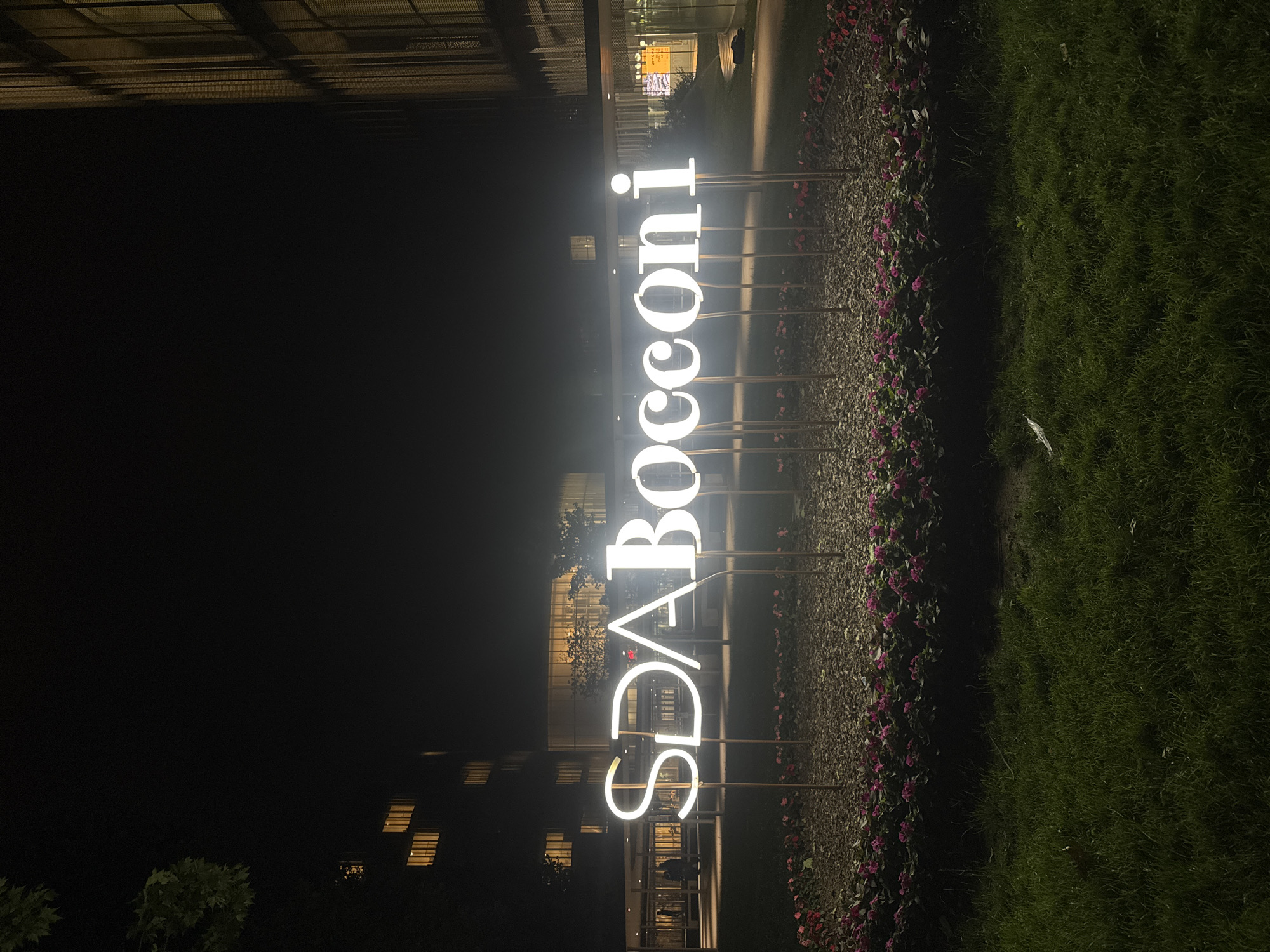 The image shows an illuminated "SDA Bocconi" sign at night in Milan, Italy. The sign is positioned in front of a building, lined with flower beds on one side and a grassy area on the other. The environment is dark, with some light visible from building windows and street lighting. There are no people visible in the immediate foreground.