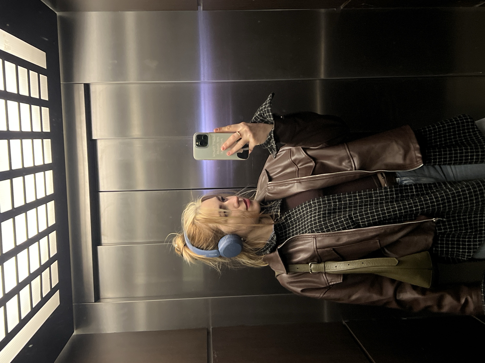 A selfie captured in an elevator features a young woman wearing blue headphones and a brown leather jacket over a plaid shirt. She is holding a phone with a case in her right hand, its screen facing away. The elevator's interior is characterized by metallic paneling and a grid of illuminated panels on one side. The location information (Boreti, Montenegro) cannot be confirmed or denied based on the image's content.