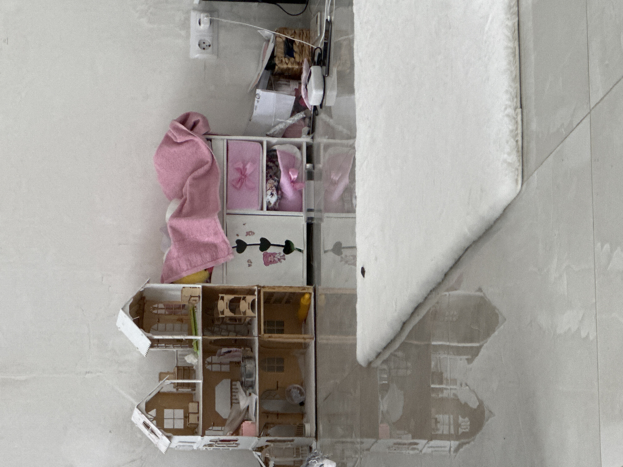 The image shows a dollhouse, a storage unit with shelves, a pink towel, and a corner of a white rug on a tile floor. The scene appears to be indoors, possibly a child's room. The location is unknown.