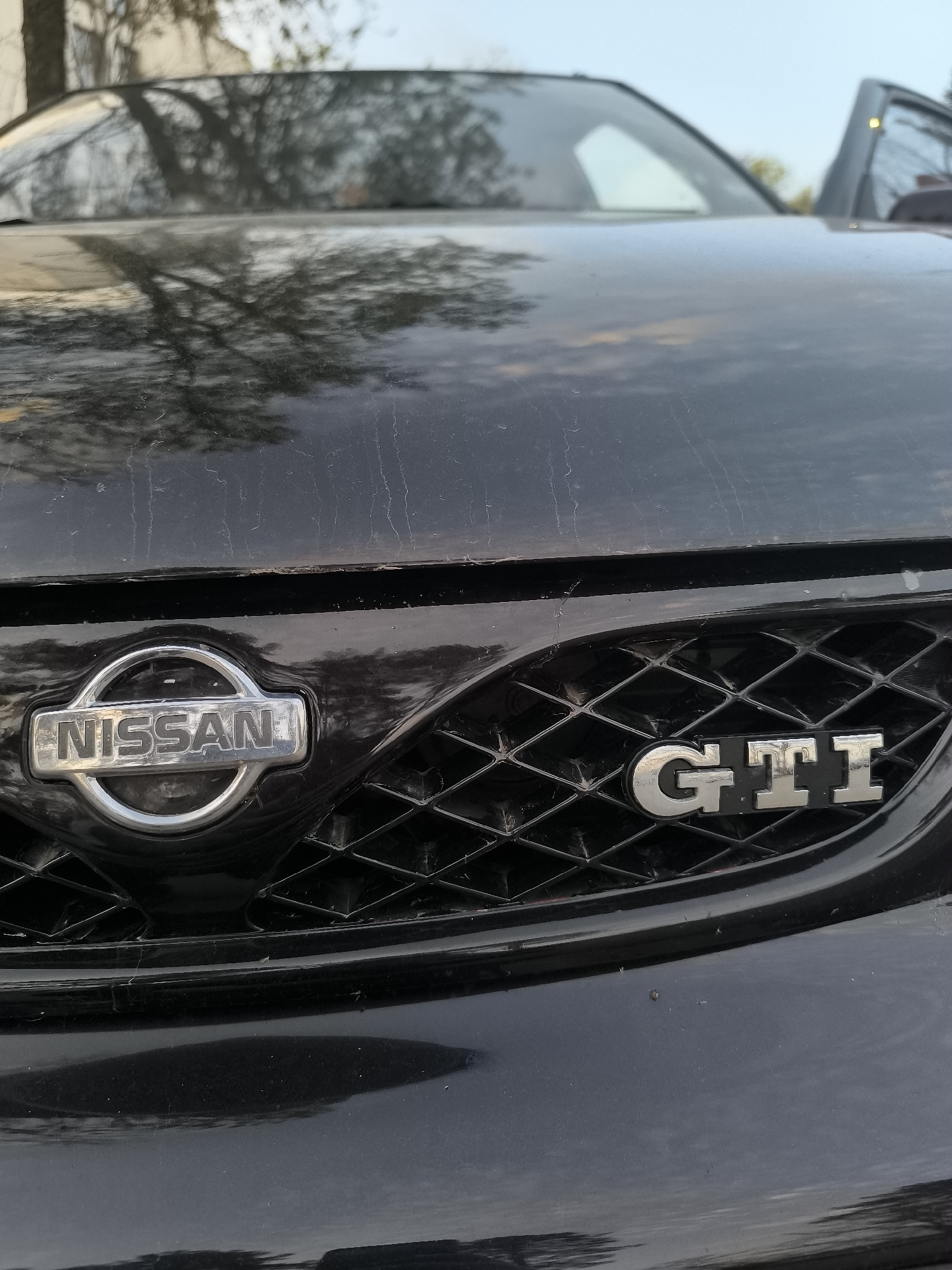 The image shows the front of a black Nissan car, specifically the grill. The Nissan logo is visible on the left, and a "GTI" badge is on the right. The hood of the car is dirty, with visible watermarks and imperfections. The reflections on the hood suggest an outdoor setting with trees and sky. There is no information that relates the location to Veliko Tarnovo, Bulgaria.