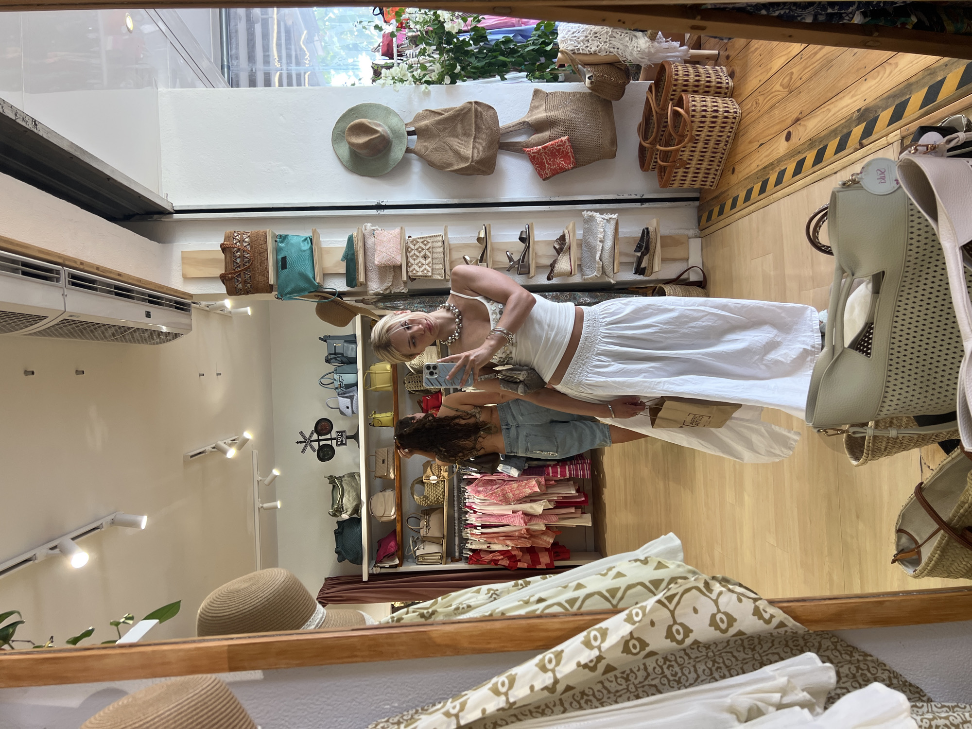 The image depicts the interior of a boutique in Pollença, Spain, likely specializing in women's clothing and accessories. In the foreground, a woman with blonde hair is taking a mirror selfie. She is wearing a white crop top and a long white skirt, accessorized with a pearl necklace and bracelets. She holds a smartphone in her right hand and a shopping bag in her left. In the mirror's reflection, another woman with dark curly hair is visible, wearing a bikini top and denim shorts, browsing a rack of clothing.
The boutique's interior is filled with merchandise. Racks of clothing, primarily in light colors and natural fabrics, are displayed. Shelves are laden with various bags, including woven straw bags, leather handbags, and tote bags, in a range of colors and textures. Hats are also prominently featured, hung on walls and displayed on shelves. The flooring is made of light-colored wooden planks. The overall ambiance suggests a summery, relaxed atmosphere, typical of a boutique in a Mediterranean tourist destination.