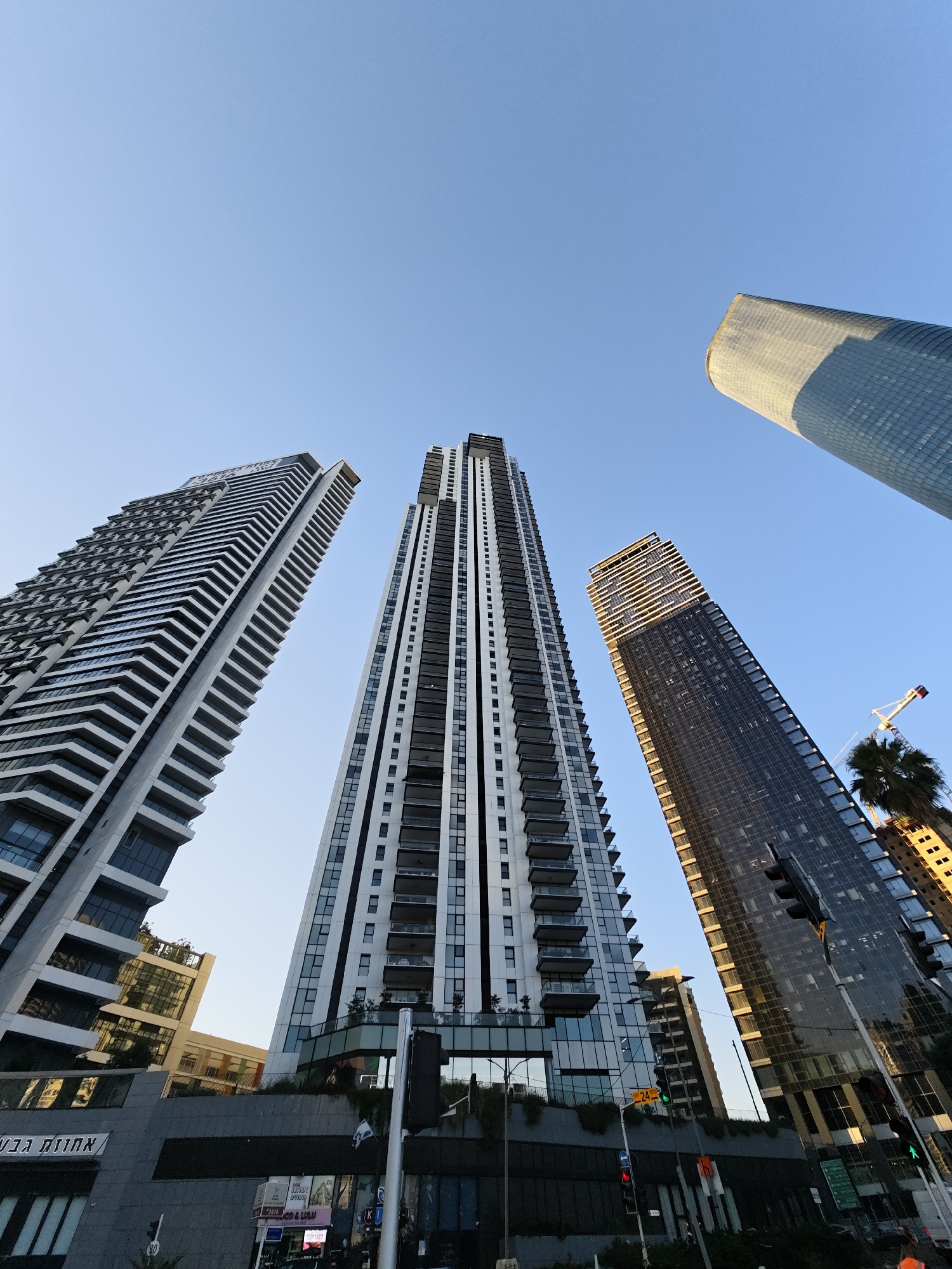This is a low-angle, wide-shot photograph of several modern skyscrapers in Givatayim, Israel. The sky is a clear, bright blue, indicating daytime. The buildings are diverse in their design, with one appearing as a cylindrical structure with a glass facade, another with a patterned facade of white and grey, a third featuring a dark, reflective glass exterior, and a fourth characterized by a stepped, terraced design. Shadows are cast on the buildings, suggesting the sun is to the right. In the foreground, a traffic light is visible, along with some street signage and a palm tree. A construction crane can be seen in the upper right portion of the image. The scene depicts an urban environment with tall, contemporary architecture.