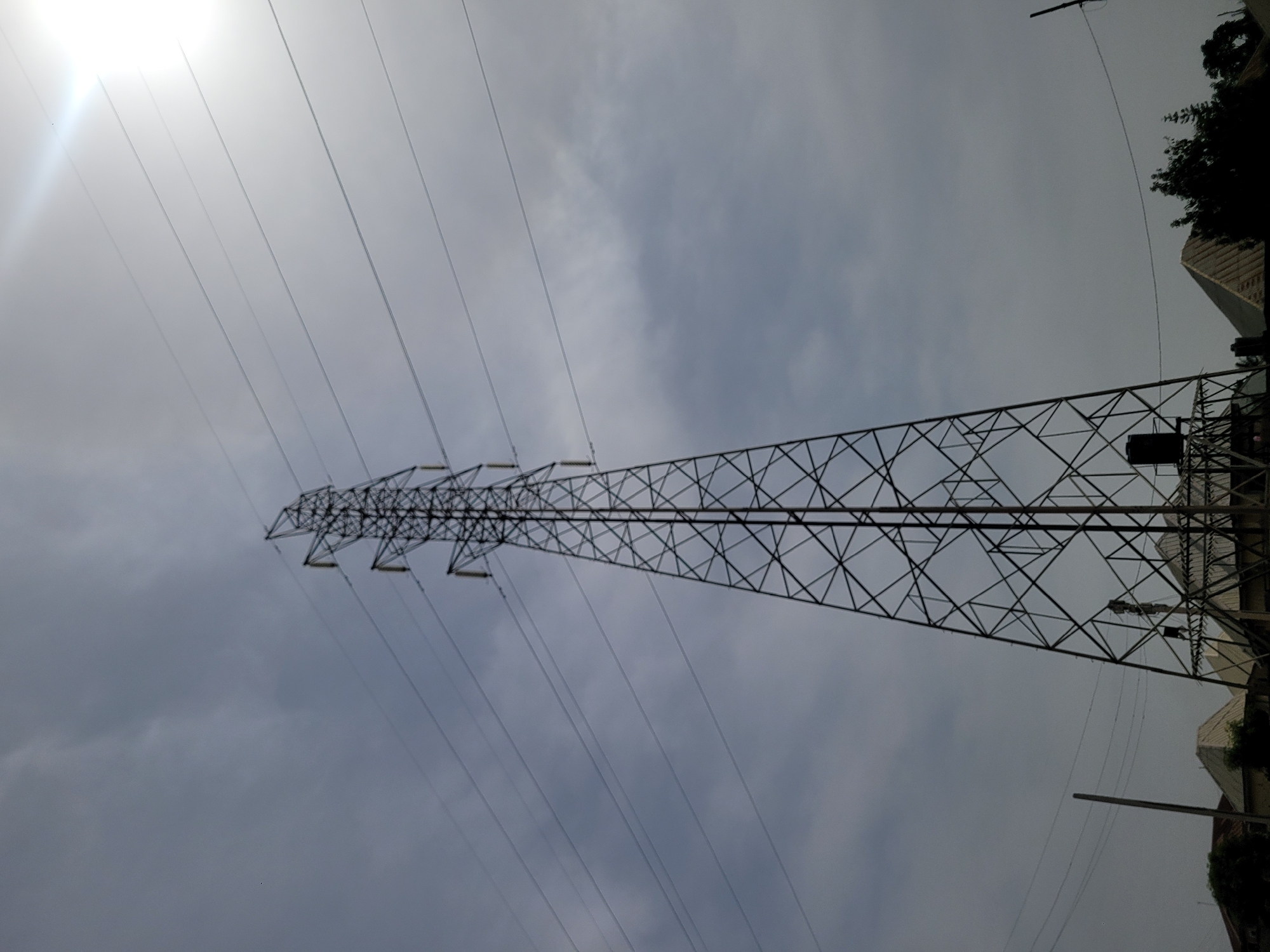 The image depicts an electrical transmission tower against a cloudy sky. The tower is located in an urban environment in Barakin Akawo, Nigeria, as evidenced by buildings and trees visible to the right of the tower. The tower itself is constructed of metal lattice and supports a series of high-voltage power lines stretching across the frame. The image is taken from a low angle, looking up at the tower. The sun is obscured by clouds in the upper left corner, creating a bright spot in the sky.