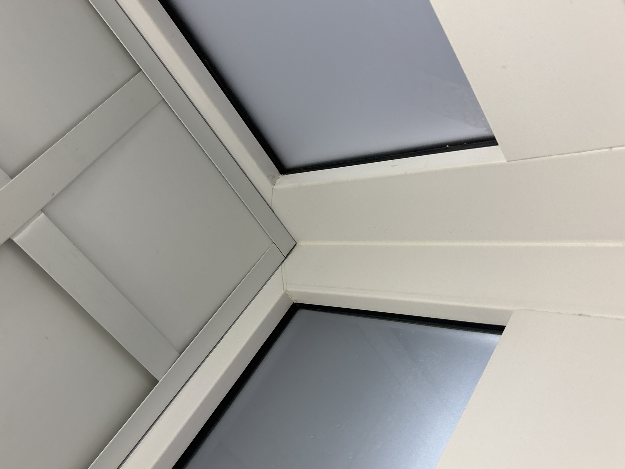 The image shows a close-up view of a white window frame intersecting with a white paneled wall. Two sections of glass are visible within the window frame, appearing gray. The paneled wall features a diagonal pattern of white panels. The location in the city of Perm, Russia is not discernible from the image.