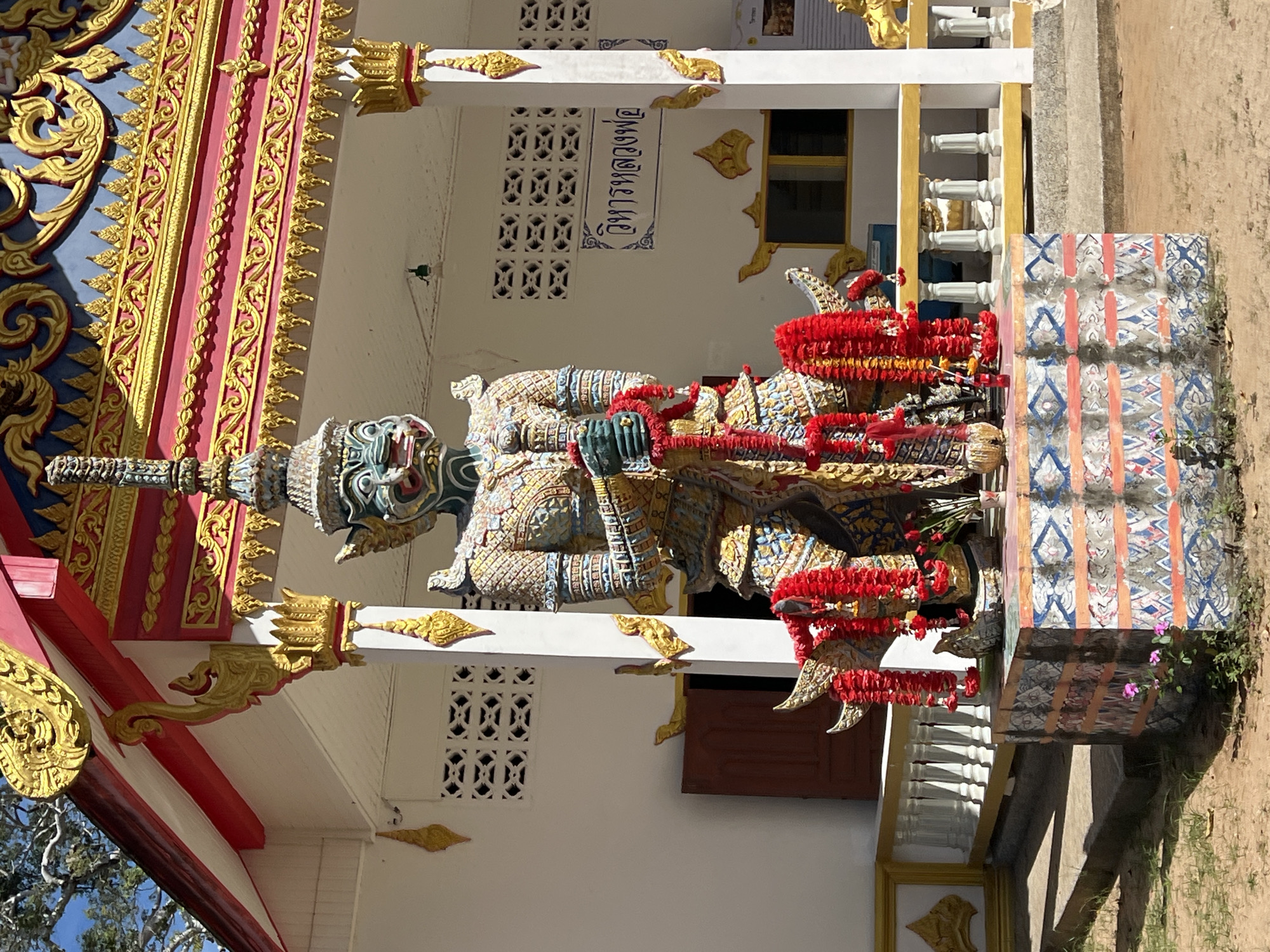 This is a full, eye-level, outdoor shot of a large, colorful statue of a mythical creature, likely a guardian from Thai folklore, positioned in front of a white building with ornate gold and red trim, typical of a temple or religious structure. The statue is imposing, with intricate details in its armor and headdress, and is adorned with strings of red flowers. It stands on a decorated pedestal. The building in the background features decorative vents and a sign with Thai script. The surrounding area is unpaved, with some grass and dirt, suggesting a rural or semi-rural setting. The overall scene captures a moment of cultural and religious significance in Baan Harn, Thailand.