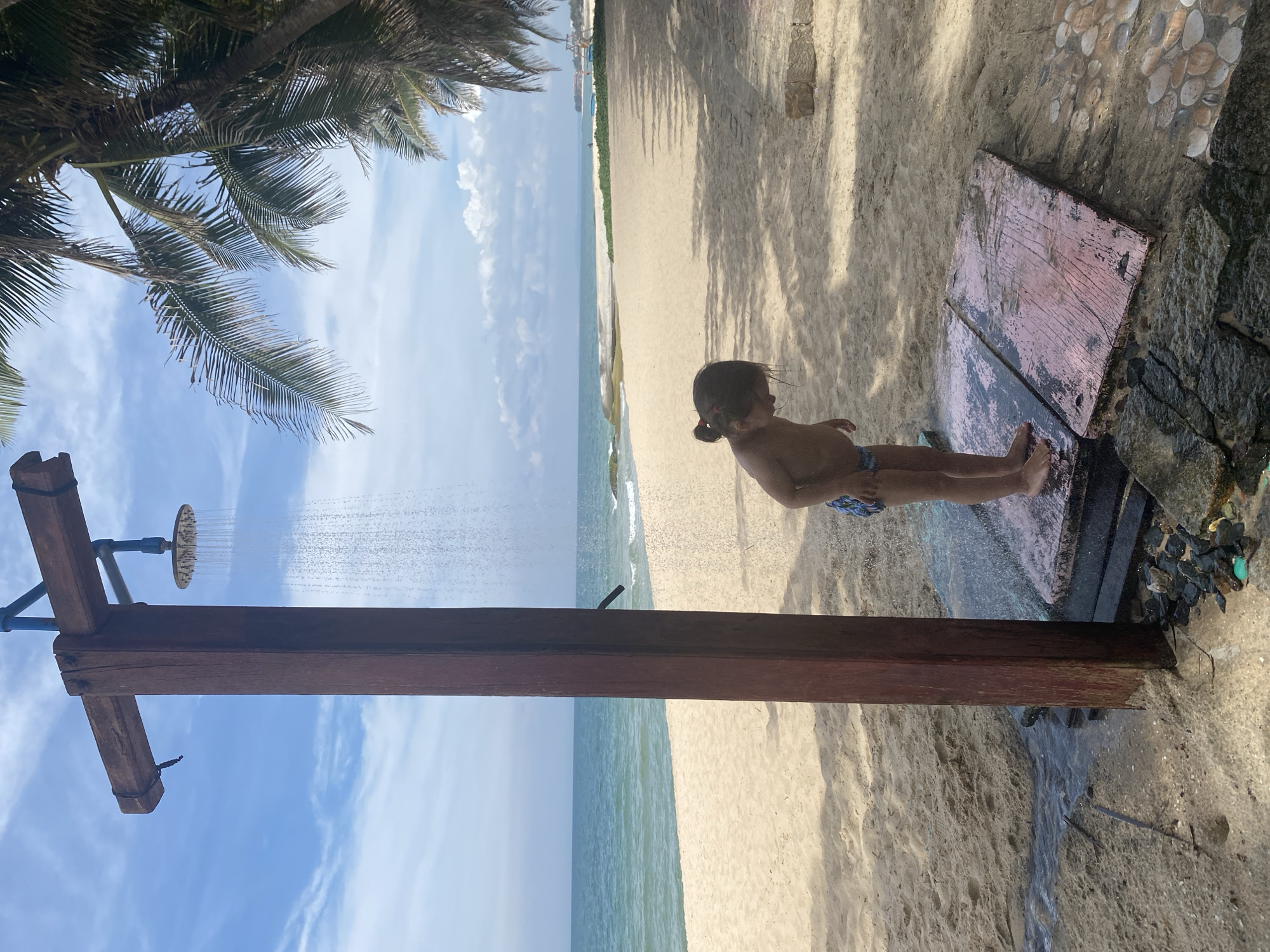 An outdoor shower structure, made of wood, stands on a sandy beach near the ocean. Water is running from the showerhead, creating a curtain of droplets. A young child, wearing patterned swim shorts, stands on a wooden platform beneath the shower, looking down. The beach is wide and sandy, with palm trees casting shadows from the right side of the frame. The ocean is visible in the background, with gentle waves breaking on the shore. The sky is blue with scattered clouds. This scene is located on the beach in Phu Thuy Ward, Vietnam.