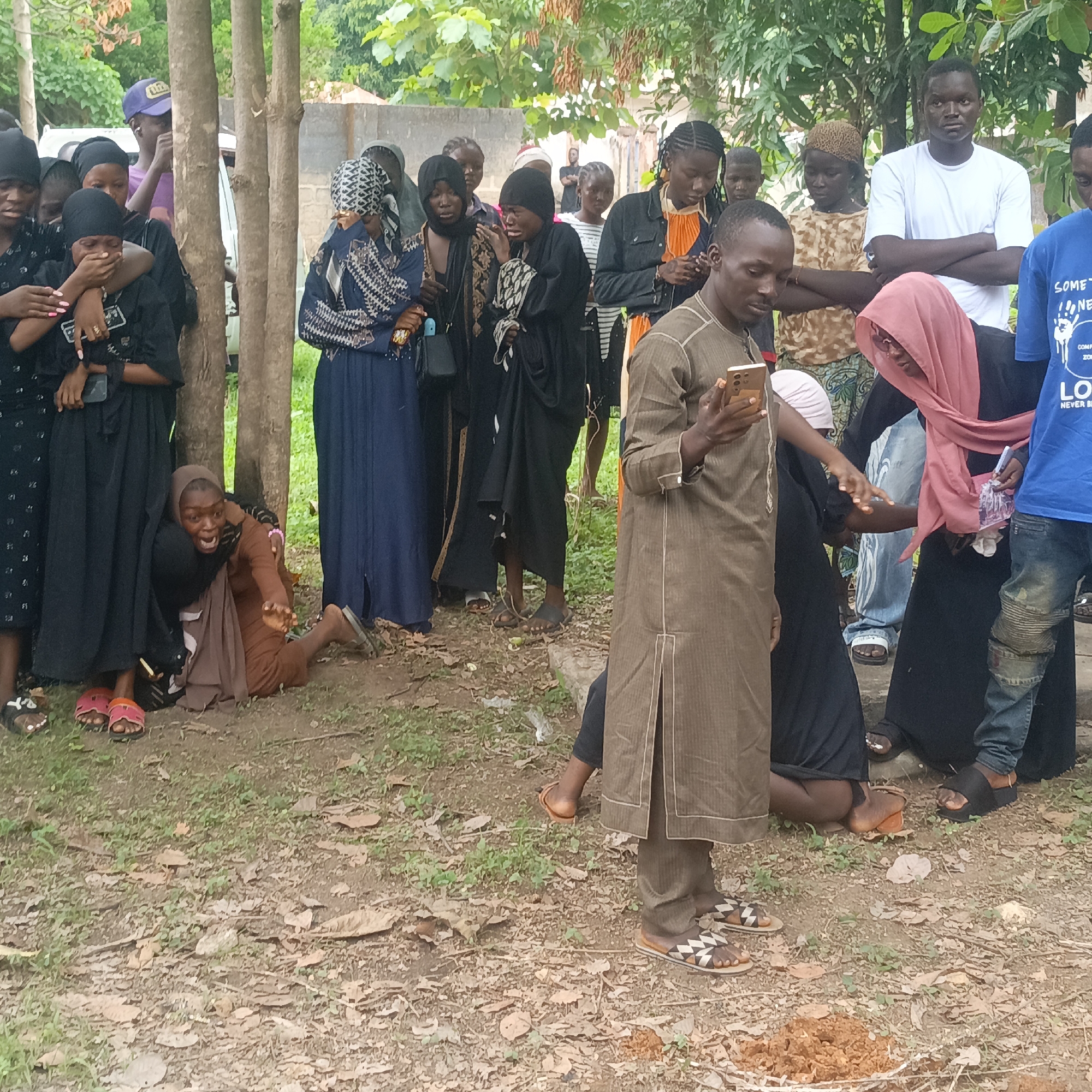 The image depicts a group of people gathered outdoors, possibly in Andaha, Nigeria. Most of the individuals appear to be in mourning, with several women visibly upset and being comforted by others. The mourners are dressed in varying shades of black and dark-colored clothing. One woman is on the ground, being held back by another person. A man in a brown robe is standing and looking at his cell phone. The environment appears to be a grassy or dirt area with trees and a building in the background. There is a small mound of dirt in the foreground.