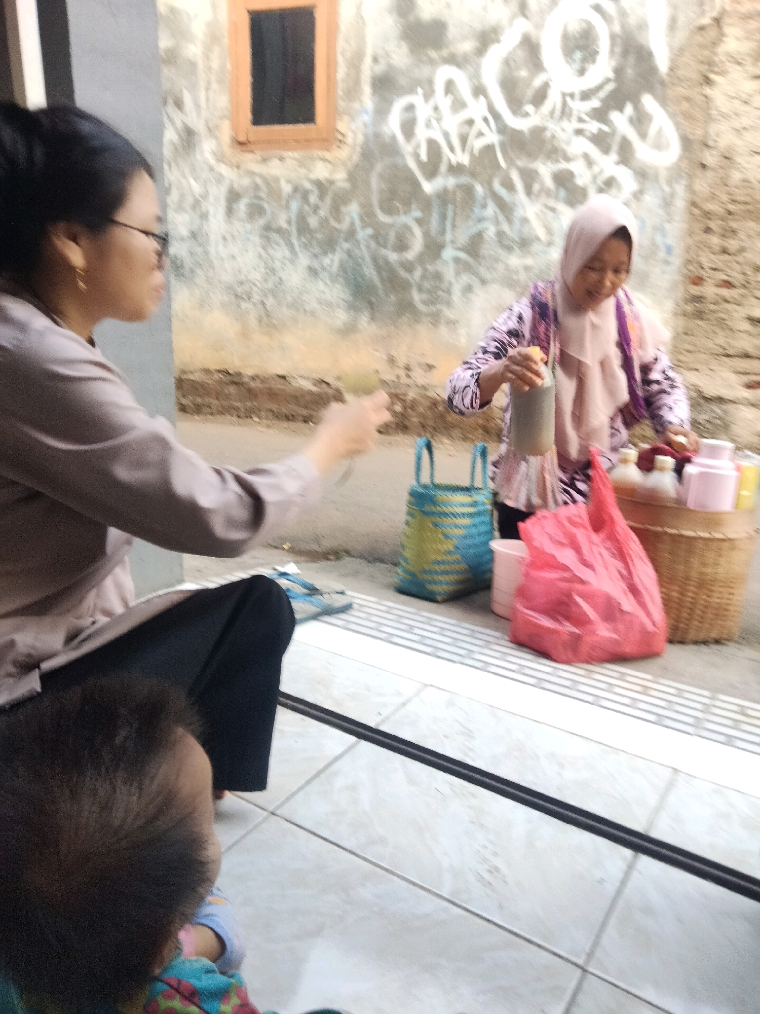 The image depicts a street scene, likely in Suka Sari, Indonesia, with two women and a baby. In the foreground, the back of a baby's head is visible. To the left, a woman wearing glasses and a light brown shirt is seated, facing away from the camera. To the right, another woman, wearing a pink hijab and a colorful patterned top, is standing. She is holding a bottle and appears to be serving a drink. Several items are placed on the ground and on a woven basket, including a bright red plastic bag, a blue and yellow woven bag, and various bottles and containers, some of which are pink. The background shows a textured wall with graffiti and a small window. The ground is tiled, with a black line running across it. The overall setting appears to be an outdoor vendor area.