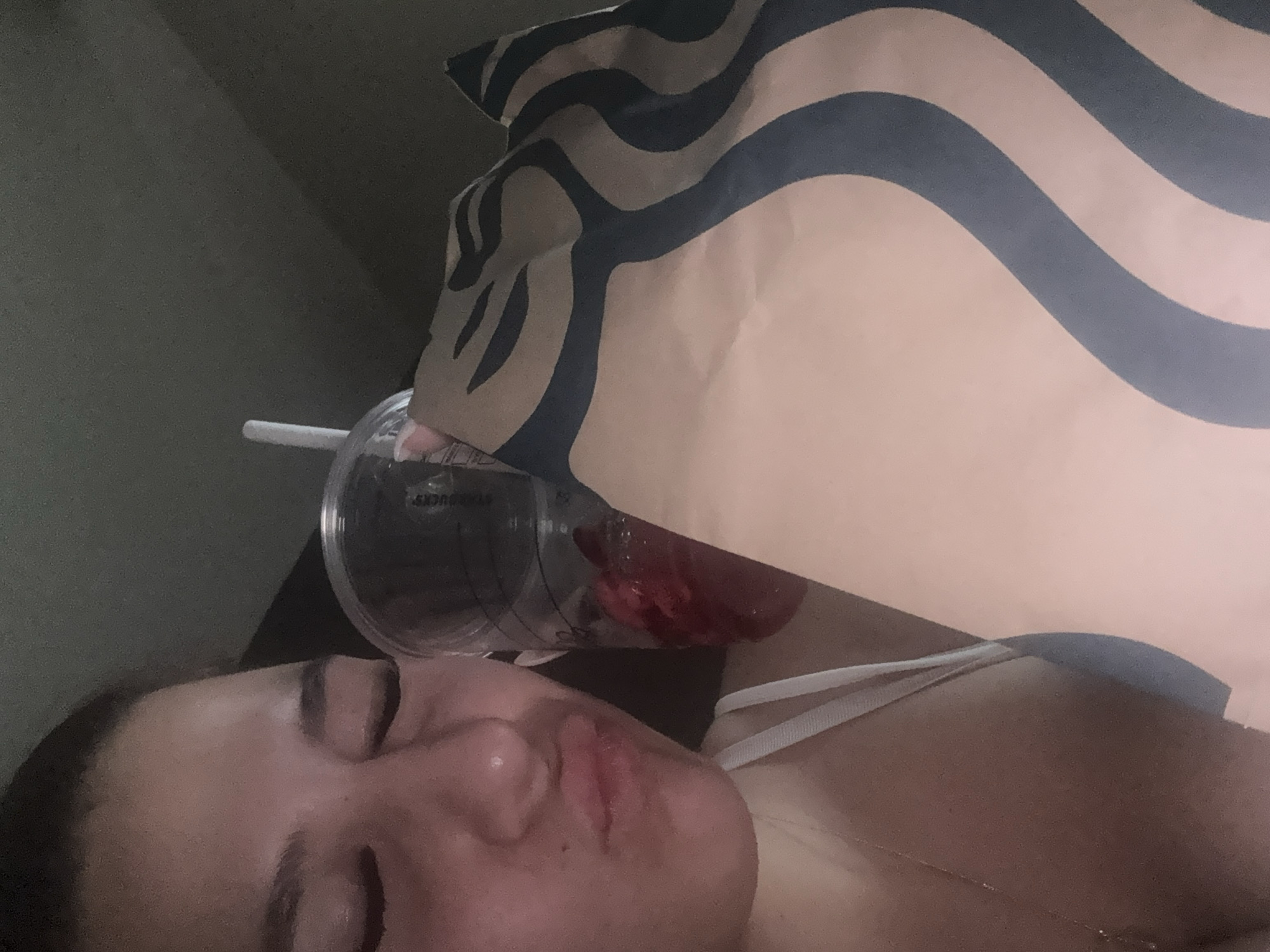 A person is depicted lying down indoors, viewed from a slightly upward angle, with their eyes closed or resting. Their head is turned slightly to the right, showing their profile. A Starbucks bag, characterized by its distinctive brown color and black wavy pattern, rests near their head, partially obscuring the view of the background. A clear plastic cup with a white straw is positioned beside the bag, also near the person's head. Inside the cup, a reddish liquid or fruit pieces are visible. The person appears to be wearing a white top with a thin strap. The lighting is dim, suggesting an indoor setting or a low-light environment. Faint text "STARBUCKS" can be seen on the rim of the plastic cup. The overall scene conveys a relaxed or resting mood.
