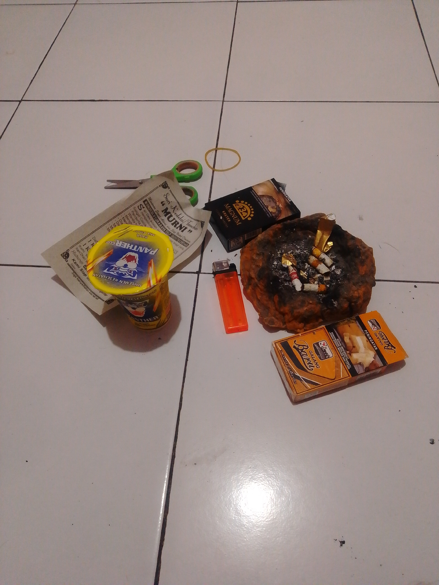 A high-angle, medium shot captures a collection of items arranged on a tiled floor in what appears to be a domestic setting in Bohar, Indonesia. The scene includes an ashtray filled with cigarette butts and ashes, a bright orange lighter, two packs of cigarettes, a cup with a yellow lid displaying a panther logo, a small pair of scissors, a piece of paper with Indonesian text, and a rubber band. The floor tiles are white with dark grout lines, and the lighting suggests an indoor environment, possibly at night. The arrangement of objects indicates a casual use of the space, with the ashtray suggesting recent smoking activity. There are no people visible in the image.