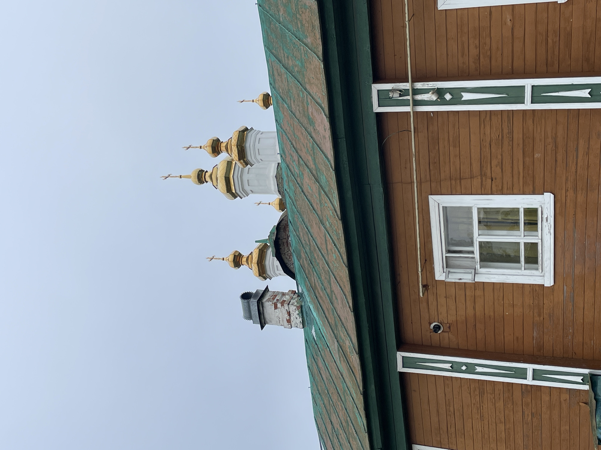 The image shows an exterior view of a wooden building with a green metal roof in Kstining, Russia. The building's facade is constructed from horizontal brown wooden planks, adorned with vertical green and white decorative elements at the corners. A white-framed window with visible internal shutters is centered on the visible portion of the wall. Above the building, the golden domes of several Orthodox churches are visible against a pale, overcast sky. A brick chimney with a metal cap protrudes from the green roof. The scene is overcast and appears to be a daytime shot. There are no people or animals present in the image.