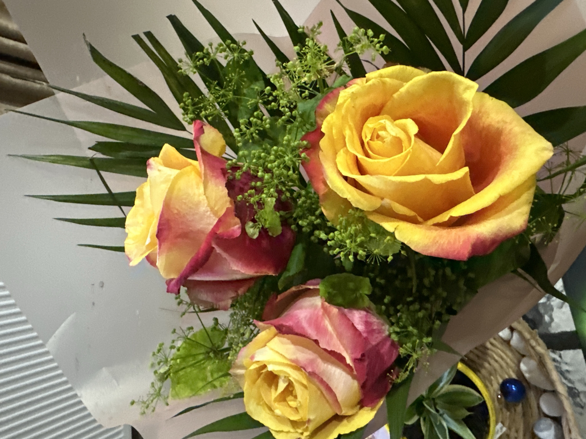 The image shows a bouquet of three two-toned roses, primarily yellow with pink/red edges. The roses are accompanied by feathery green leaves and small green umbel-like clusters. The bouquet is wrapped in light-colored paper. In the lower right corner, part of a wicker basket with white stones and a yellow pot with a plant are visible. The location is not specified in the image.