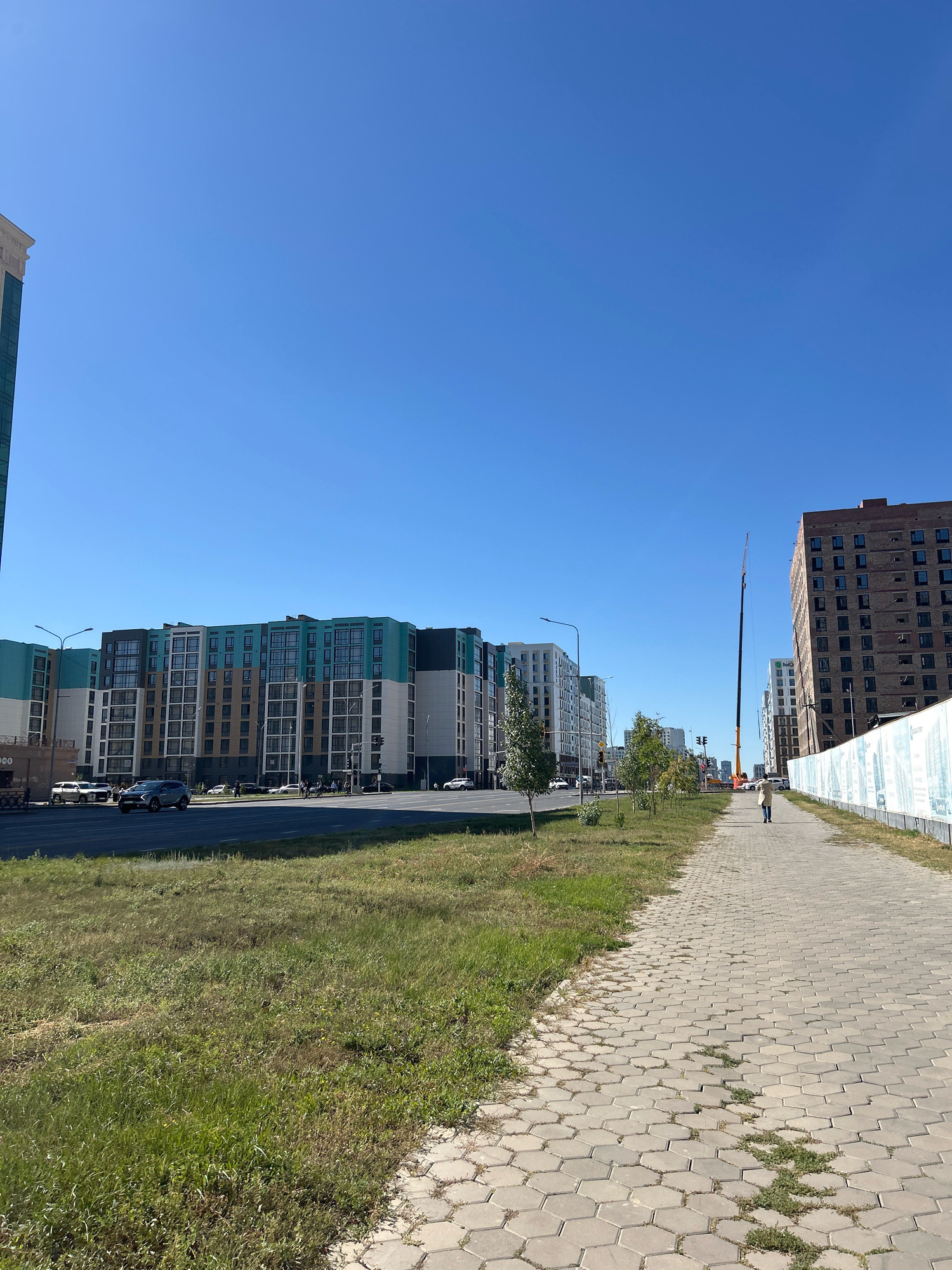 A street scene in Astana, Kazakhstan, features a modern residential complex with a turquoise and brown facade on the left, opposite a multi-story brick building under construction on the right. A tall construction crane is visible between the buildings. A paved pedestrian walkway runs along the right side of the street, bordered by a grassy median. A lone individual in a light-colored coat walks away from the camera on the walkway. Several cars are driving on the multi-lane road. The sky is clear and blue.